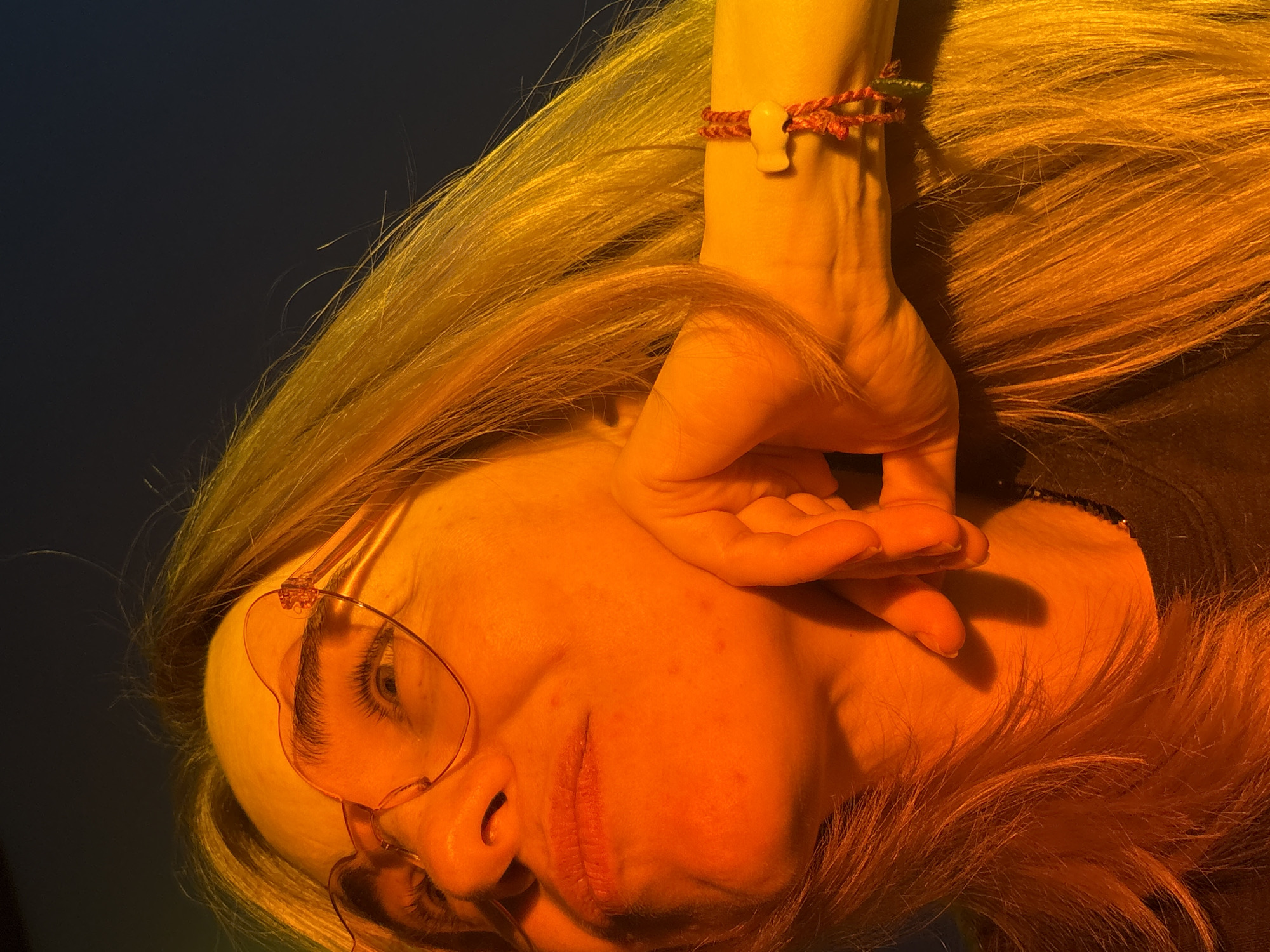 A close-up, rotated portrait features a person with long, flowing blonde or light brown hair, illuminated by intense, warm golden-orange light against a dark, indistinct background. The image is rotated 90 degrees counter-clockwise, making the subject's head appear on the left and their hair cascading upwards.
The subject's face, likely female, is partially visible, with one eye looking towards the viewer through clear or light-tinted glasses with distinctive frames. A hand, with fingers gently curled, rests near the face. A red braided bracelet with a small, off-white, stylized charm is worn on the wrist. The strong, directional light highlights the texture of the hair and skin, emphasizing subtle details like small blemishes on the cheek and forehead. The lips are visible, appearing neutral or slightly parted. The overall impression is an artful, stylized pose under dramatic lighting. There is no visible text.