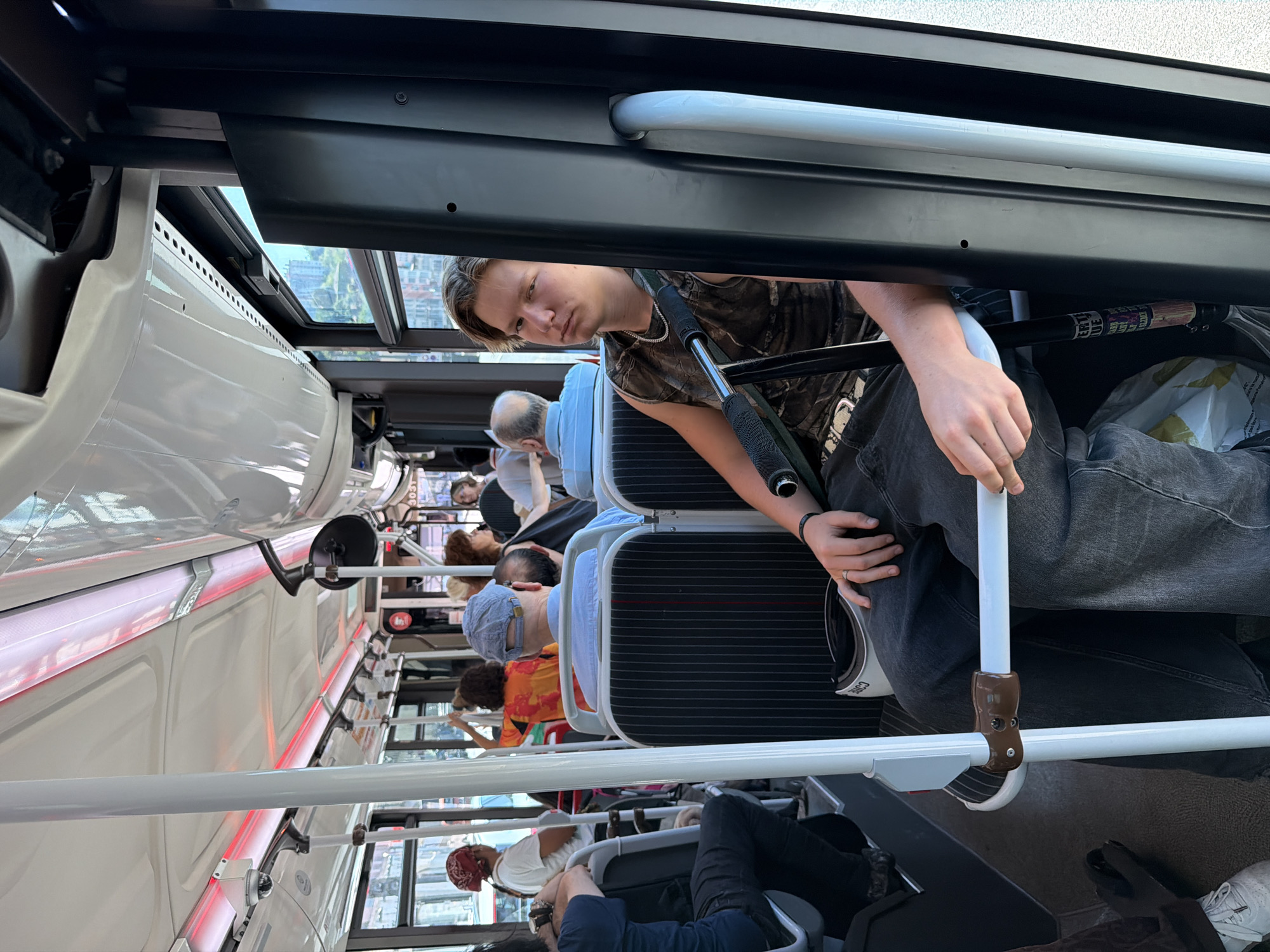 The image shows the interior of a public bus. A young man with blond hair, wearing a camouflage-patterned tank top and dark jeans, is seated on the right side of the bus, facing forward and slightly to the left. He is leaning back against a pole and has his right arm resting on a metal bar. His gaze is directed towards the camera. Other passengers are visible in the background, some seated and some standing. The bus has white overhead panels with red lighting strips. Windows line the left side of the bus, offering a view of buildings and a cityscape outside. The scene appears to be in Lyon, France, given the presence of a bus with this specific interior design, commonly found in the city's public transportation system, and the urban architecture visible through the windows. The overall atmosphere is ordinary, depicting a typical scene on a public bus.
