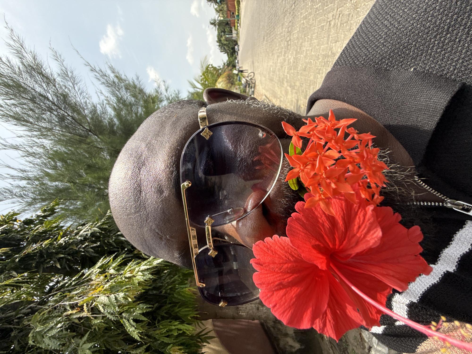 The media file presents a close-up, upward-angled shot of an individual, seemingly a selfie, taken outdoors in bright daylight. The subject is a person with dark skin, a bald head, and visible stubble or short facial hair on their chin and jawline. They are wearing large, dark-lensed sunglasses with gold-toned frames. A distinctive gold Louis Vuitton monogram, a four-lobed star, is visible on the temple of the sunglasses. Faint text, "Louis VUIT," can also be seen on the bottom left lens.
Two types of vibrant flowers are prominently featured near the person's right ear (viewer's left). There is a large, fully bloomed red hibiscus flower with its long pistil extending outwards, and a cluster of smaller, star-shaped orange flowers, likely Ixora. These flowers appear to be tucked into or held against the side of the person's head.
The individual is wearing a dark-colored garment, possibly a sweater or jacket, which features a textured, knitted pattern and a visible silver zipper pull on the right side. A white, possibly striped or zig-zag, pattern is visible on the lower part of this garment.
In the background, under a bright sky with scattered white clouds, there are green trees and foliage, including what appears to be a casuarina (pine-like) tree and broad-leafed plants. A paved road or path is also visible, leading towards distant buildings and what looks like a bicycle. The strong sunlight casts clear shadows, suggesting the photo was taken during late morning or early afternoon. The setting is an outdoor, possibly urban or suburban environment, consistent with Lekki, Nigeria.