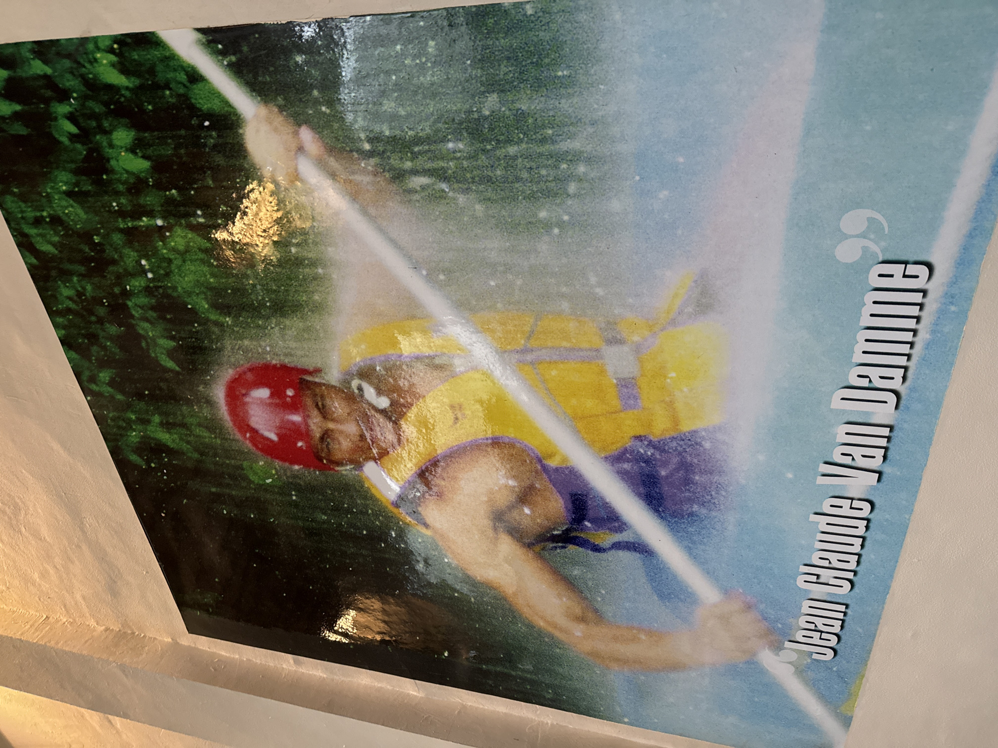 The image is a photograph of a poster featuring Jean Claude Van Damme, appearing to be a still from a movie or an action shot. He is depicted in a white kayak, wearing a red helmet and a yellow life vest over a purple garment. Water splashes around him, suggesting he is engaged in whitewater kayaking or a similar water sport. He is holding a white paddle and his expression is intense. The background consists of lush green foliage, likely trees, and the water is a vibrant blue. Text on the poster reads "Jean Claude Van Damme". The poster is mounted on a textured, light-colored wall, possibly made of concrete or stucco. The lighting in the scene is somewhat diffused, with some reflections visible on the glossy surface of the poster. There is no specific indication of its location in Petang, Indonesia, beyond it being a poster displayed in a public or private space.
