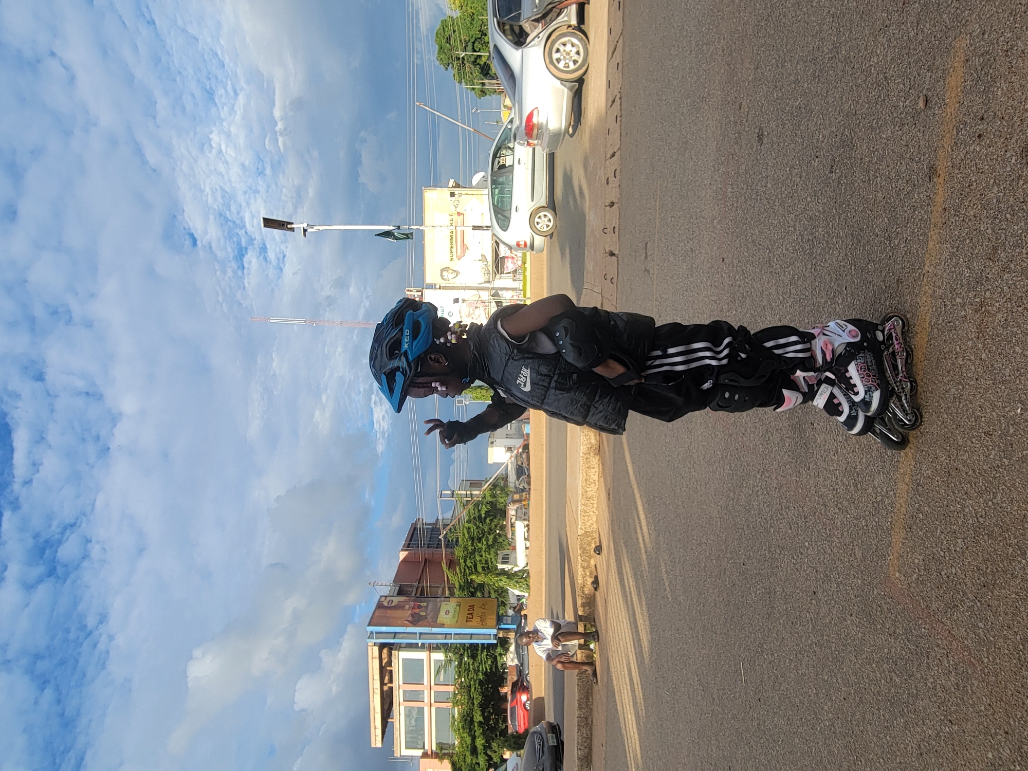 An outdoor, eye-level full shot in Jos, Nigeria, depicts a young child in the foreground standing on an asphalt road while wearing full inline skating gear. The child, with braided hair, wears a blue and black KED helmet, a black Nike-branded padded vest (with additional Korean text), black track pants with three white stripes, black elbow and knee pads, and black and pink inline rollerblades. The child's right hand is raised with two fingers extended in a V-sign.
In the mid-ground to the left, an adult male wearing a white shirt and light-colored shorts sits on a concrete curb next to his removed slippers. Behind him, a silver sedan is parked. Further back, a multi-story building with a light-colored facade and a large billboard featuring "TEA DA" text is visible, partially obscured by green trees.
In the mid-ground to the right, two silver vehicles, a hatchback and a sedan, are on the road. A white pole with a solar-powered street light extends upwards. Behind it, a white sign with "SUPERMARKET" text and another sign featuring a man's portrait are visible. A red and white communication tower is present in the far left background. The sky above is partially cloudy with blue patches and white clouds. Faint shadows are cast on the road.
