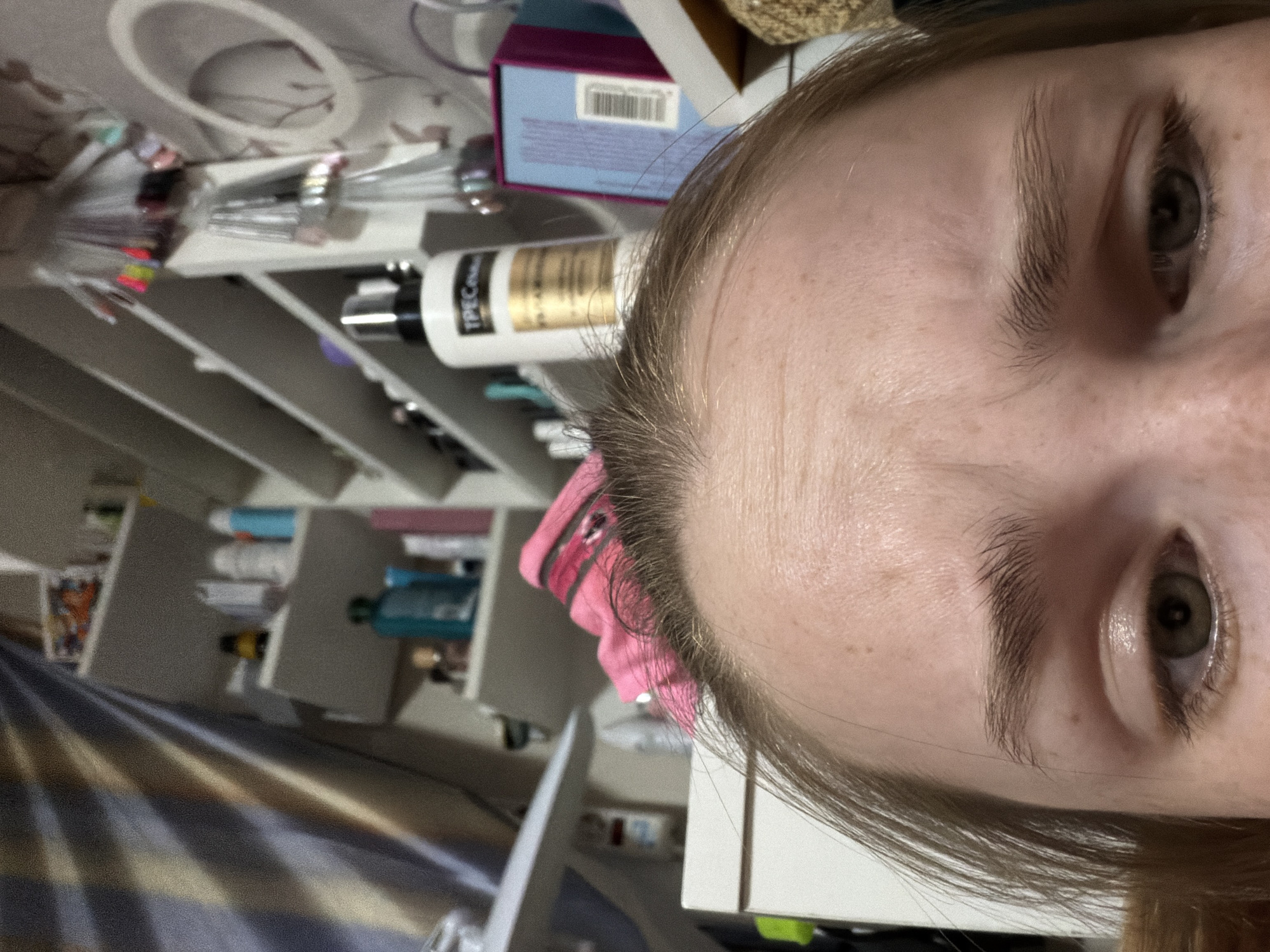 A close-up, angled image shows the face of a person with light skin, visible eyebrows, and eyes. The background features a shelving unit filled with various items, including containers and bottles of beauty products, nail polish, and some boxes. The overall scene appears to be in a private indoor space, likely a bedroom or bathroom. The image does not contain sufficient information to determine if it was taken in Berezniki, Russia or any other specific location.