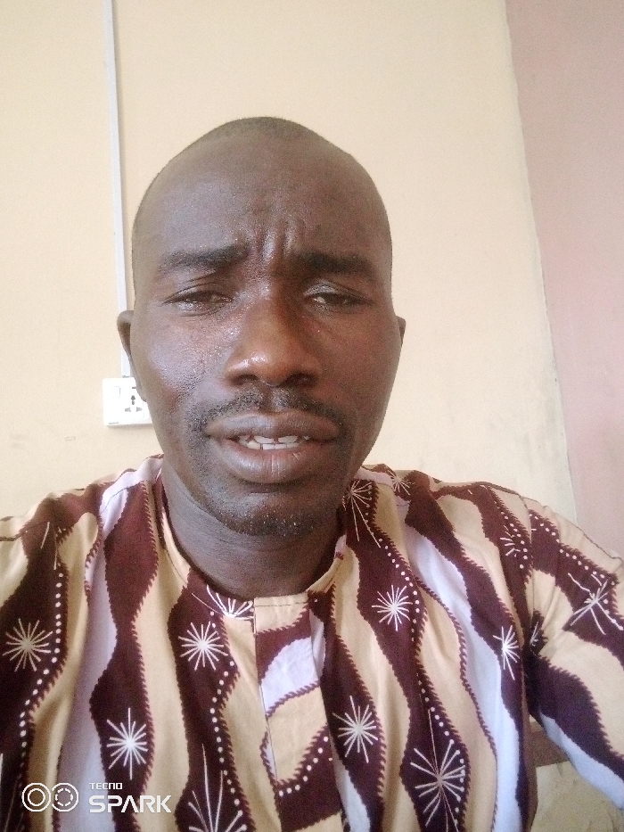 A close-up portrait shows an adult male of African descent, positioned in an indoor setting within Ajaokuta, Nigeria. The subject has a short, dark hairstyle, a moustache, and stubble. His brow is furrowed, and his mouth is slightly open, revealing some teeth. A visible sheen is present on his forehead. He is wearing a patterned traditional garment with sections of brown, beige, and white, adorned with white starburst designs and dotted lines. The background features a plain wall, predominantly light beige on the left side and a lighter tan or pinkish hue on the right. A white two-gang electrical outlet with three-pin sockets and a white vertical conduit are visible on the upper left portion of the wall. The lower-left corner of the image displays a 'TECNO SPARK' watermark.