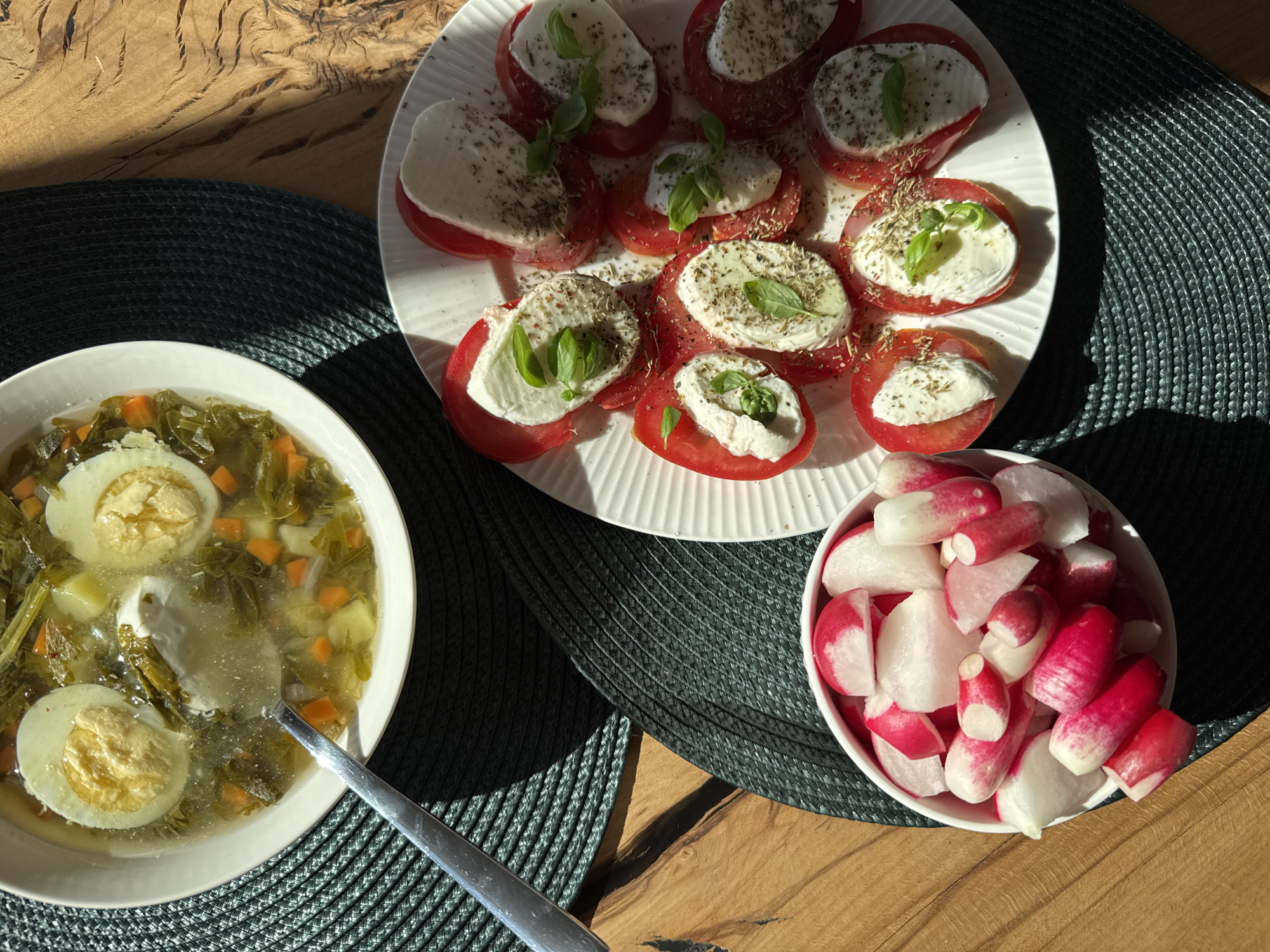 This top-down, natural light photograph captures a rustic table setting featuring a meal in what appears to be a home kitchen in Steklyannyi, Russia. The scene is set on a wooden table, with a dark, woven placemat underneath two dishes and a bowl.
The primary subjects are the food items. A white plate holds a colorful caprese salad, composed of alternating slices of ripe red tomatoes and fresh white mozzarella, each topped with a sprig of basil and a sprinkle of dried herbs. Adjacent to the caprese salad is a white bowl filled with sliced radishes, their vibrant pink and white hues contrasting with the dark placemat. To the left, a white bowl contains a hearty vegetable soup. The soup is a clear, green broth filled with leafy greens, diced carrots, potatoes, and two halves of a hard-boiled egg. A sliver of sour cream sits on top of the soup, and a silver spoon rests on the edge of the bowl.
The natural light suggests it is daytime, likely late morning or early afternoon, given the warm shadows cast across the table. The overall impression is one of a simple, healthy, and home-cooked meal. There is no visible text in the image.