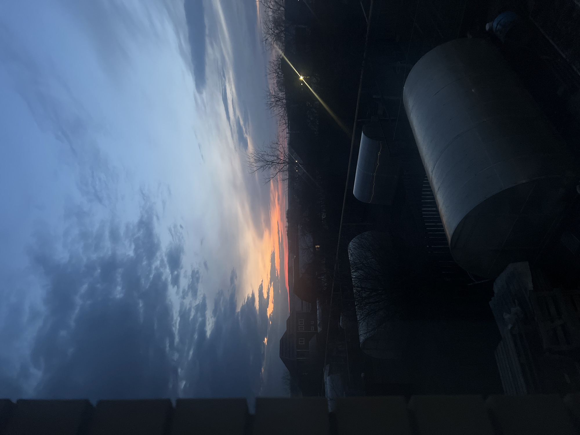 The image depicts a sunset over an urban landscape, likely taken from an elevated position like a balcony or rooftop. The sky displays a gradient of colors, transitioning from dark blue with clouds on the left to orange and pink hues near the horizon. Silhouetted buildings are visible against the sunset. To the right, a cylindrical metal tank dominates the foreground, with other smaller industrial or utility structures nearby. A sparse tree line and a street light add to the urban setting. There are no people visible in the scene. Without additional landmarks, pinpointing the precise location in Votkinsk is impossible.