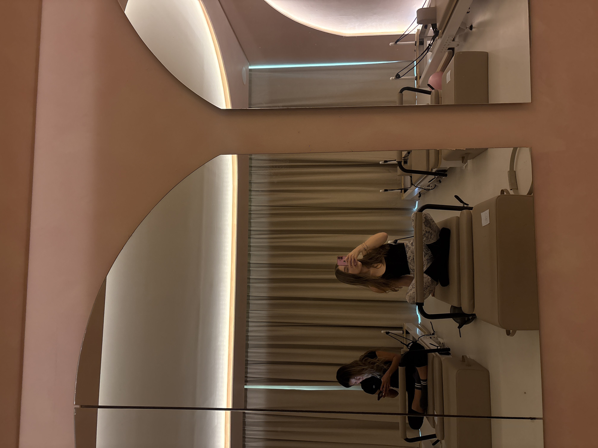 The image shows a room, likely a salon or studio, in Mexico City, Mexico. The scene is captured in two large, irregularly-shaped mirrors mounted on a light pink wall. In the lower mirror, two reflections of a woman are visible. One reflection shows her sitting on a beige chair, looking down and holding a smartphone. She is wearing a black top, black pants, and a cap. The other reflection shows the same woman holding her phone and looking at herself. The reflections show a brightly lit interior with light beige drapes and equipment behind the woman. The upper mirror reflects the same equipment and drapes, without a person present.