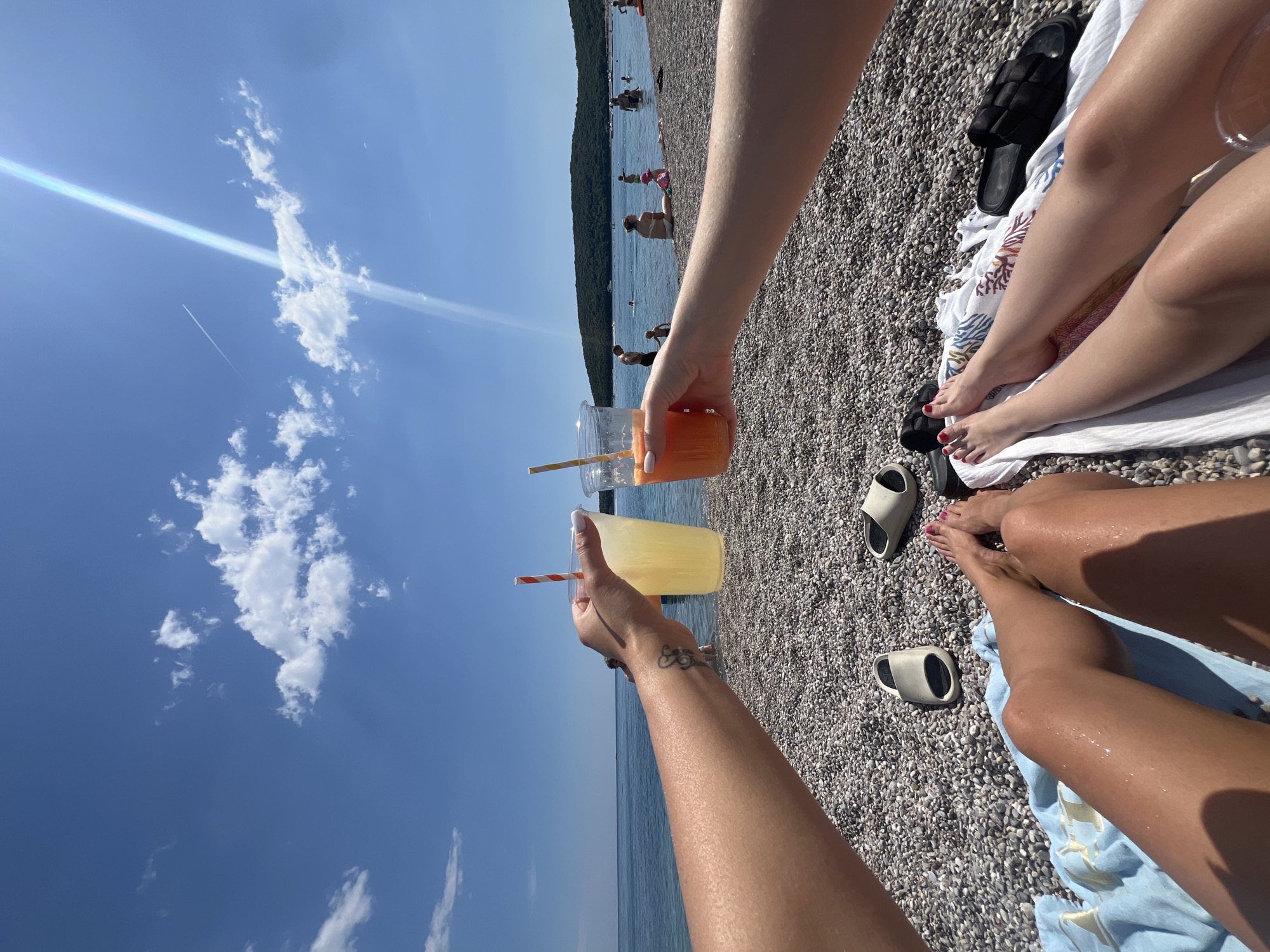 Two people sit on a pebble beach in Prijevor, Montenegro, raising their drinks towards each other in a toast. The person on the left holds a yellow drink, while the person on the right has an orange drink. The sea and blue sky stretch behind them. People are visible in the water and along the shoreline in the distance. Sandals are lying nearby on towels.