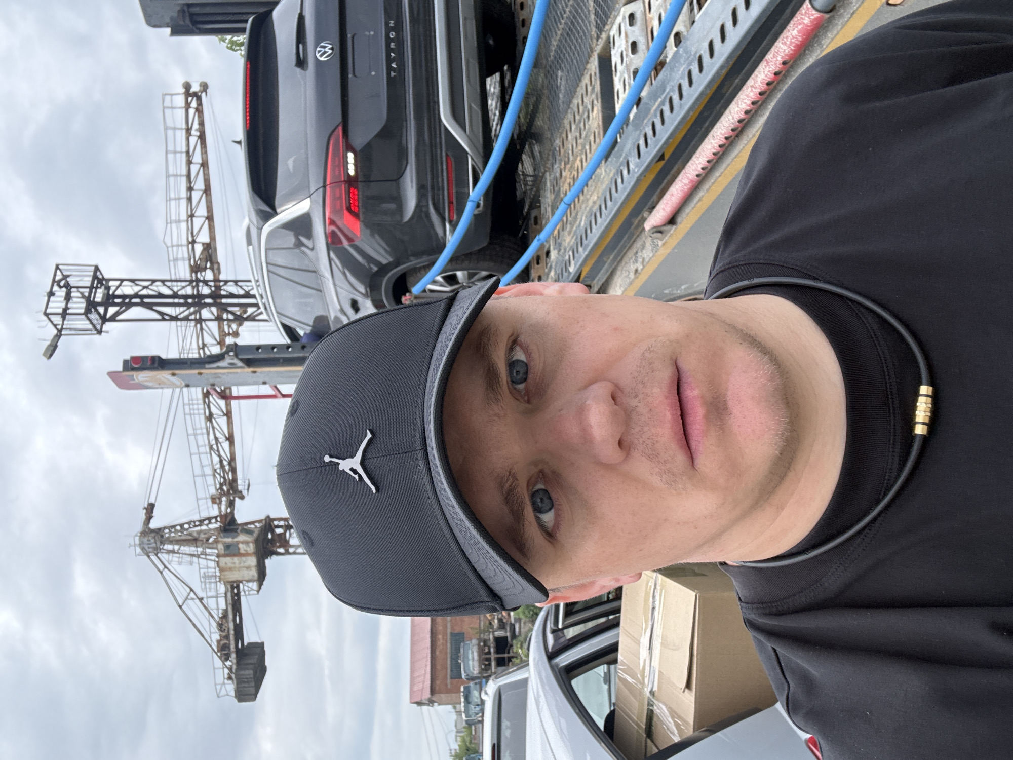 A man is taking a selfie in what appears to be an industrial or logistics setting, likely a port in Vladivostok, Russia. He has blue eyes and a neutral expression, wearing a black t-shirt and a black baseball cap with a white Jordan logo. A black neckband with a gold accent is visible around his neck.
Behind him, a black Volkswagen Tayron SUV is loaded onto a car carrier or trailer, with blue straps/hoses visible along the carrier's side. The rear of the SUV displays the "TAYRON" model name and the Volkswagen emblem. In the background, under an overcast sky, a large, rusty industrial crane structure is prominent. Partially visible in the lower right are cardboard boxes in the back of a white vehicle. The overall impression is one of goods transport and industrial activity under cloudy daytime conditions.