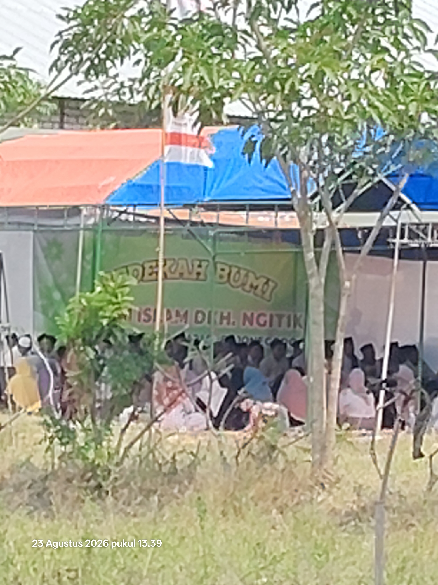 An outdoor gathering is taking place under a blue and orange tarp canopy. A green banner with white lettering is visible, reading "BERKAH BUMI" at the top and "TISIAM DIH. NGITIK" below. A group of people are seated and standing in front of the banner, some wearing religious headwear like peci and hijabs. The environment appears to be grassy and somewhat overgrown. The date and time stamp "23 Agustus 2026 pukul 13.39" is superimposed in the lower left corner, suggesting the image was taken in Padang Mentoyo, Indonesia.