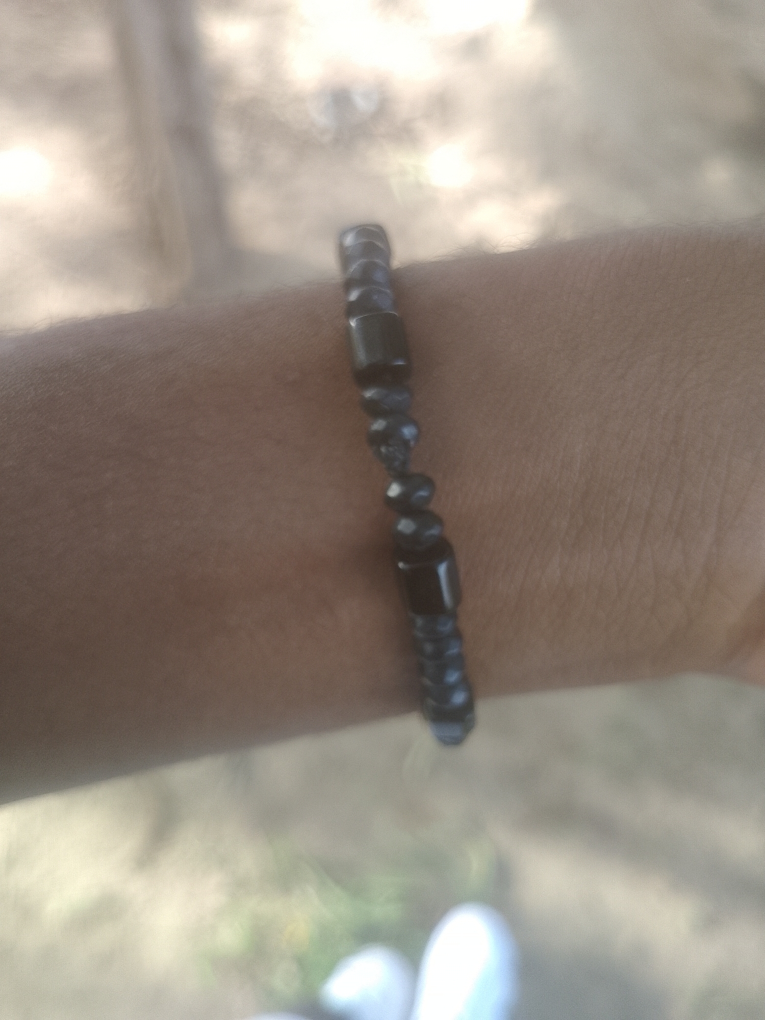 The image is a close-up of a person's arm wearing a beaded bracelet. The bracelet is made of dark, possibly black, beads, some of which are cylindrical and others round. The arm is tanned and appears to be hairless. The background is blurred and out of focus, suggesting an outdoor setting with soft lighting. The ground in the background appears to be dirt or sand, with some hints of green vegetation and lighter colored objects that might be footwear. The overall impression is a casual, everyday moment captured. There is no visible indication of the city of Majemu, Nigeria in the image itself.