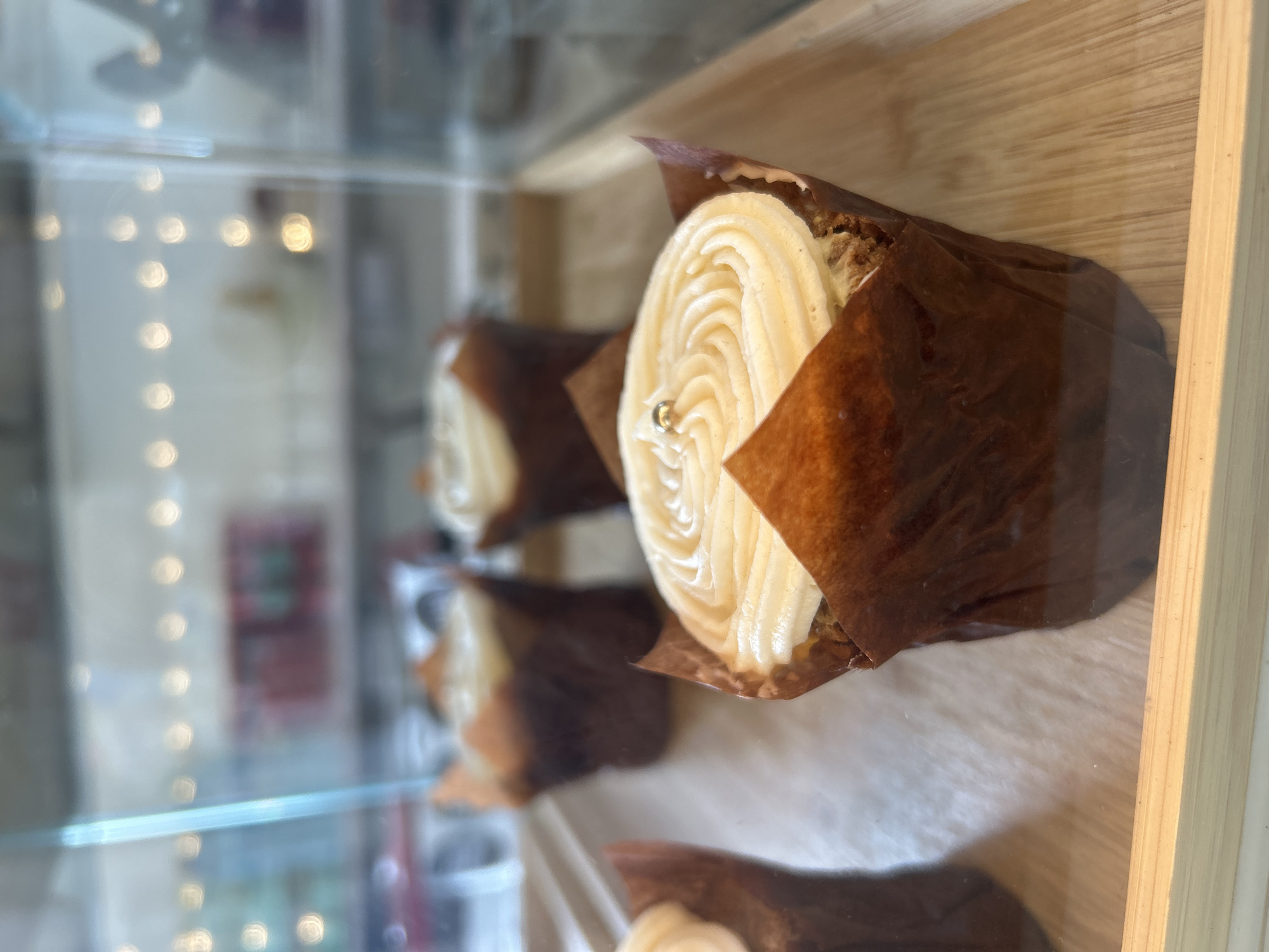 The image captures a close-up view of several cupcakes displayed inside a glass case, seen from a slightly elevated and angled perspective. The primary subject is a cupcake in the foreground, adorned with a tall, light-colored, creamy frosting piped in an elegant swirling pattern. A small, shiny silver bead or sprinkle decorates the peak of the frosting. This cupcake is cradled in a dark brown, crinkled paper liner.
Behind the leading cupcake, at least two more identical cupcakes are visible in a row, receding into soft focus. They share the same light frosting and dark brown liners, suggesting a consistent product line. The cupcakes rest on a light-toned wooden surface, possibly a shelf or tray, which features a subtle wood grain pattern.
Reflections and out-of-focus circular lights (bokeh) are visible on the glass of the display case and in the background, indicating an indoor setting with artificial lighting, typical of a bakery, cafe, or dessert shop. The overall atmosphere suggests a presentation of fresh, appealing baked goods. No visible text is present in the image.
