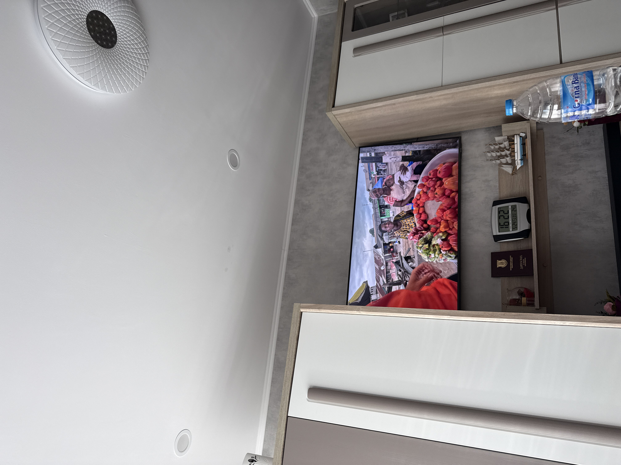 The image shows an interior setting, likely a residential or hotel room, in Lovech, Bulgaria. A ceiling light fixture is affixed to the white ceiling. A modern television is mounted on the wall. The television screen displays a scene from a foreign market, possibly in Africa, where people are selling produce. Beneath the TV, there is a small shelf holding a digital clock displaying the time as 16:22. Next to it, a shelf holding a bottle of water and a decorative ship. The TV is flanked by a white cabinet with a light brown trim. Below this, another white cabinet with a grey trim.