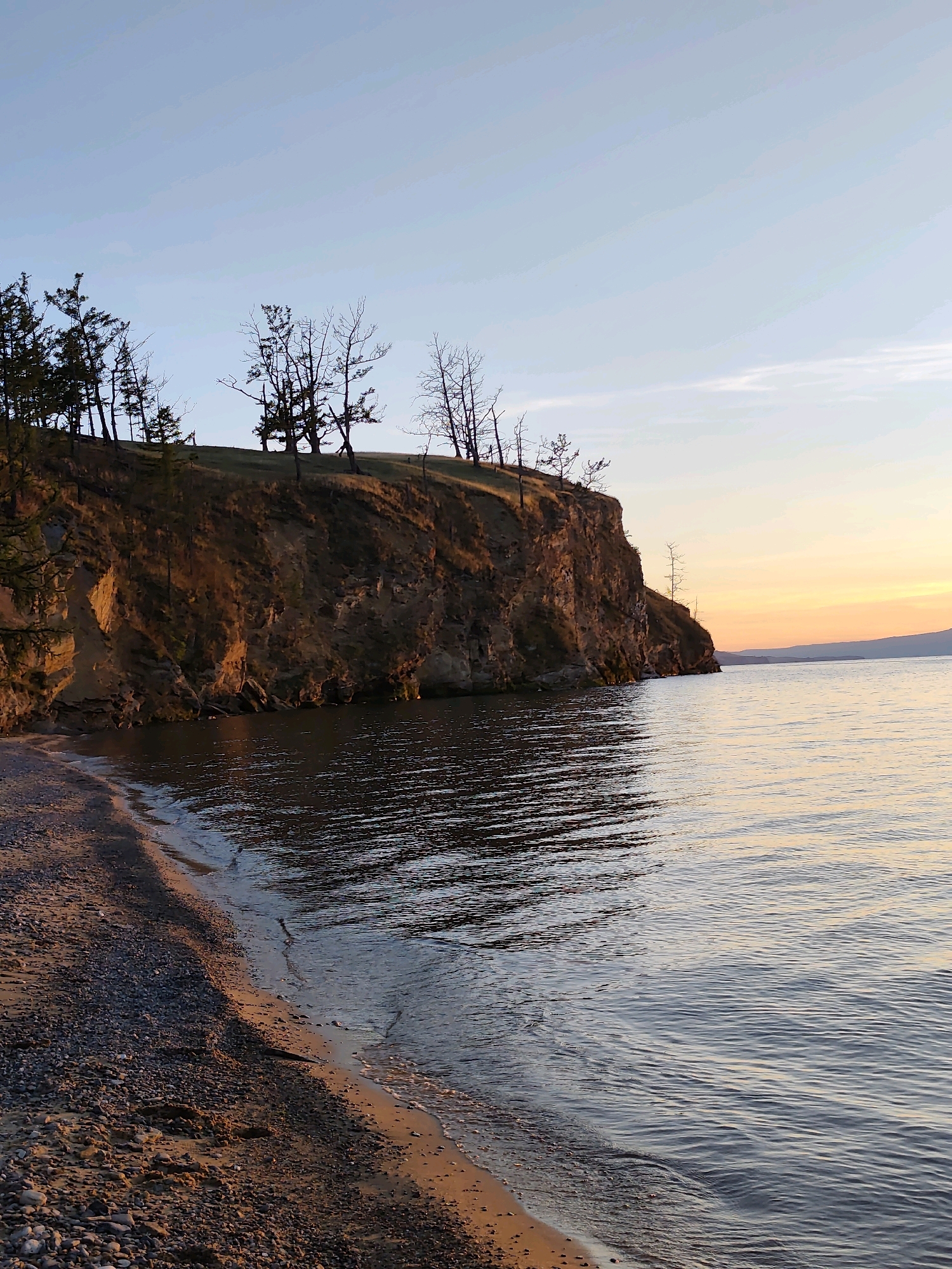 This is a vertical, medium-shot, eye-level outdoor photograph of a cliff overlooking a body of water at sunset, likely located near the village of Kharantsy, Russia. The foreground consists of a pebbly beach with a narrow strip of wet sand where small waves are gently lapping. The middle ground features a steep, rocky cliff face, illuminated by the warm, golden light of the setting sun, casting deep shadows. Sparse, bare trees are visible at the top of the cliff, silhouetted against the sky. The background is dominated by a vast expanse of calm water, reflecting the soft hues of the sunset. The sky transitions from a pale blue at the top to warm orange and pink tones near the horizon, where distant hills are faintly visible. There are no people or animals present in the image. The overall atmosphere is serene and natural.
