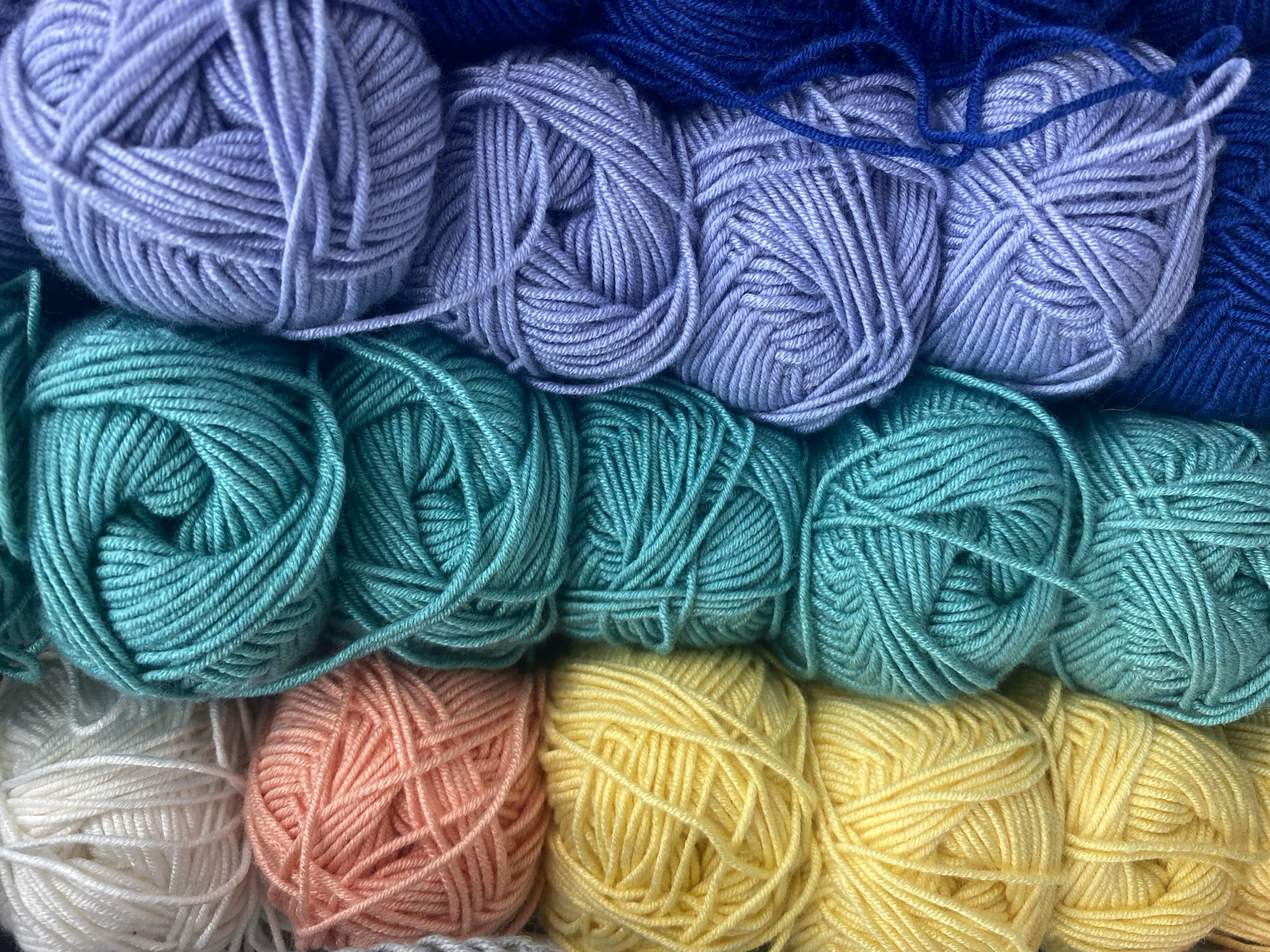 This is a close-up, overhead shot of neatly stacked balls of yarn, arranged in rows. The yarn is soft and appears to be made of acrylic or a similar synthetic material. The colors are vibrant and include white, peach, yellow, turquoise, lavender, and navy blue. The yarn is wound into loose balls, with some stray strands visible. The lighting is bright and even, highlighting the textures of the yarn. There are no people, animals, or any indications of a specific location within Krasnodar, Russia, or any other city. The image is solely focused on the yarn.