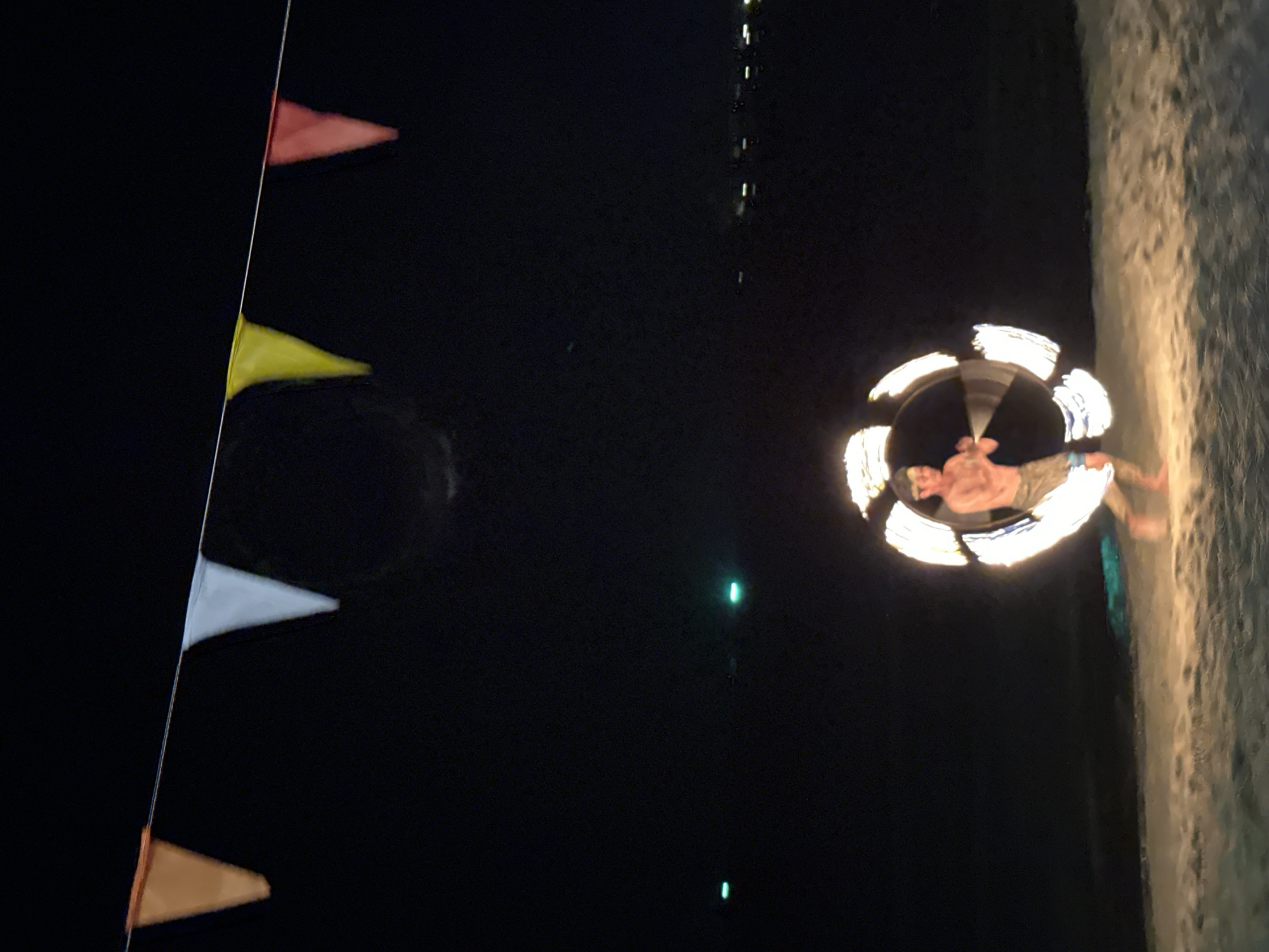 At night in Hat Khai Muk, Thailand, a shirtless man spins a circle of fire. The location appears to be a beach or sandy area. A string of triangular flags, mostly red, yellow, white, and orange, is to the left, and dim lights are in the background.