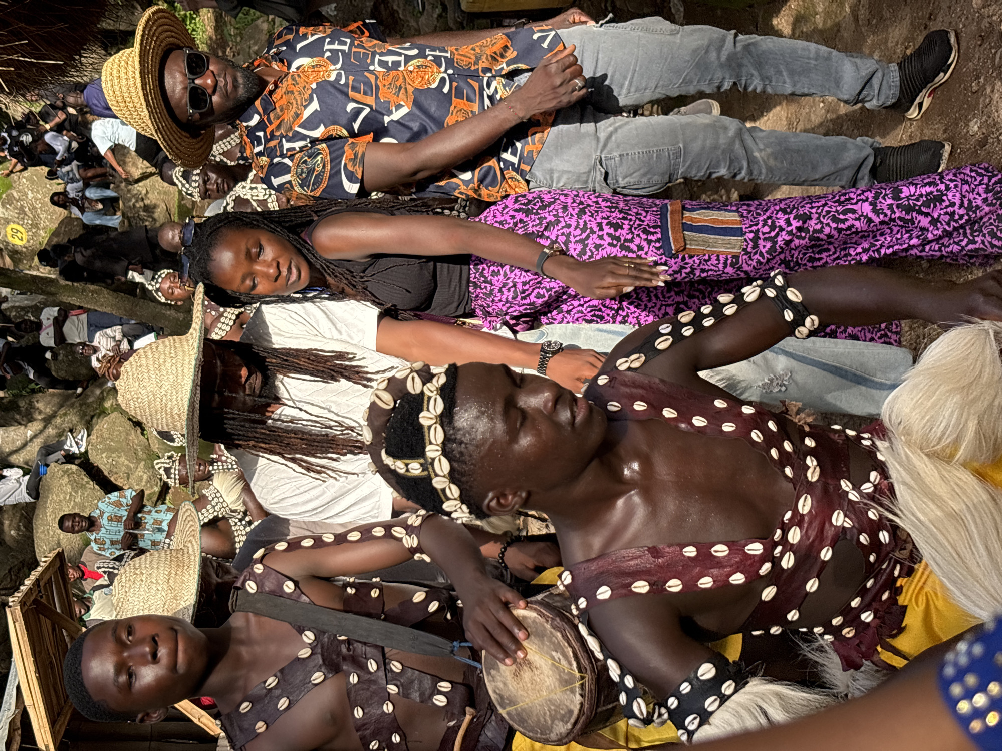 The image depicts an outdoor gathering of people, likely in a traditional or cultural context, given the attire of some individuals. In the foreground, three young men are prominently featured. The one on the left wears a straw hat and a decorative outfit adorned with numerous white shells. The central figure, also in a shell-adorned outfit, has dreadlocks and is resting his head on a woman's shoulder. He is holding a small drum and appears to be participating in an activity. The third young man, on the far right, is partially visible and is also wearing a shell-adorned garment.
Behind them, a woman with braided hair is lying down, her head resting on the shoulder of a man. The man wears a straw hat and sunglasses, and an eye-catching, brightly colored shirt with a pattern of gold and black symbols. Both are dressed in casual Western-style clothing, with the man in grey jeans. Other individuals are visible in the background, some sitting or lying on the ground, suggesting a relaxed or celebratory event. One person in the background is wearing a traditional patterned shirt.
The scene appears to be set during the day, with dappled sunlight filtering through the trees, indicating an outdoor location. The ground is rocky and uneven, and there is vegetation visible. The presence of traditional attire and the relaxed atmosphere suggest a cultural celebration or gathering. The overall mood seems to be one of community and shared experience, possibly a festival or a communal rest period. There is no visible text in the image.