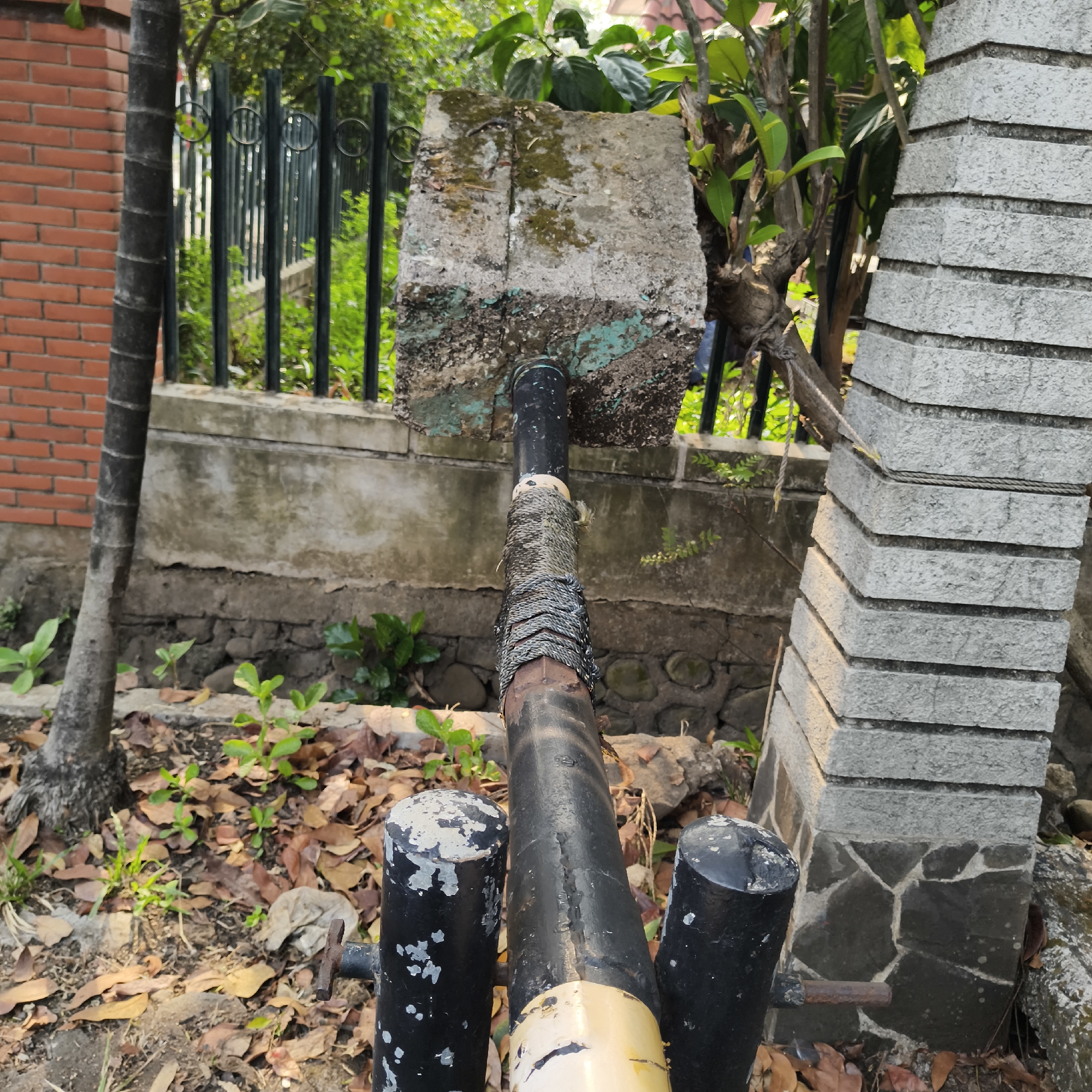 The image is an outdoor scene at a location that appears to be an exterior wall or boundary in South Jakarta, Indonesia. In the foreground, a series of black metal posts are visible, one of which is topped with a weathered white cap. Extending upwards from these posts is a black pipe, wrapped with rough, dark-colored rope for insulation or grip. This pipe is positioned against a deteriorating concrete structure, part of which is painted with faded turquoise and white. Beyond the concrete structure, a black metal fence with circular decorative elements encloses a garden area with lush green foliage. To the left of the fence, a brick wall constructed from reddish-brown bricks is visible, with a tall, dark-barked tree trunk growing next to it. To the right of the concrete structure, a pillar constructed from grey stone blocks is visible. The ground in the foreground and midground is covered with dry, fallen leaves and scattered stones, with some small green plants emerging through the debris. The overall impression is of an aged, slightly neglected outdoor space.
