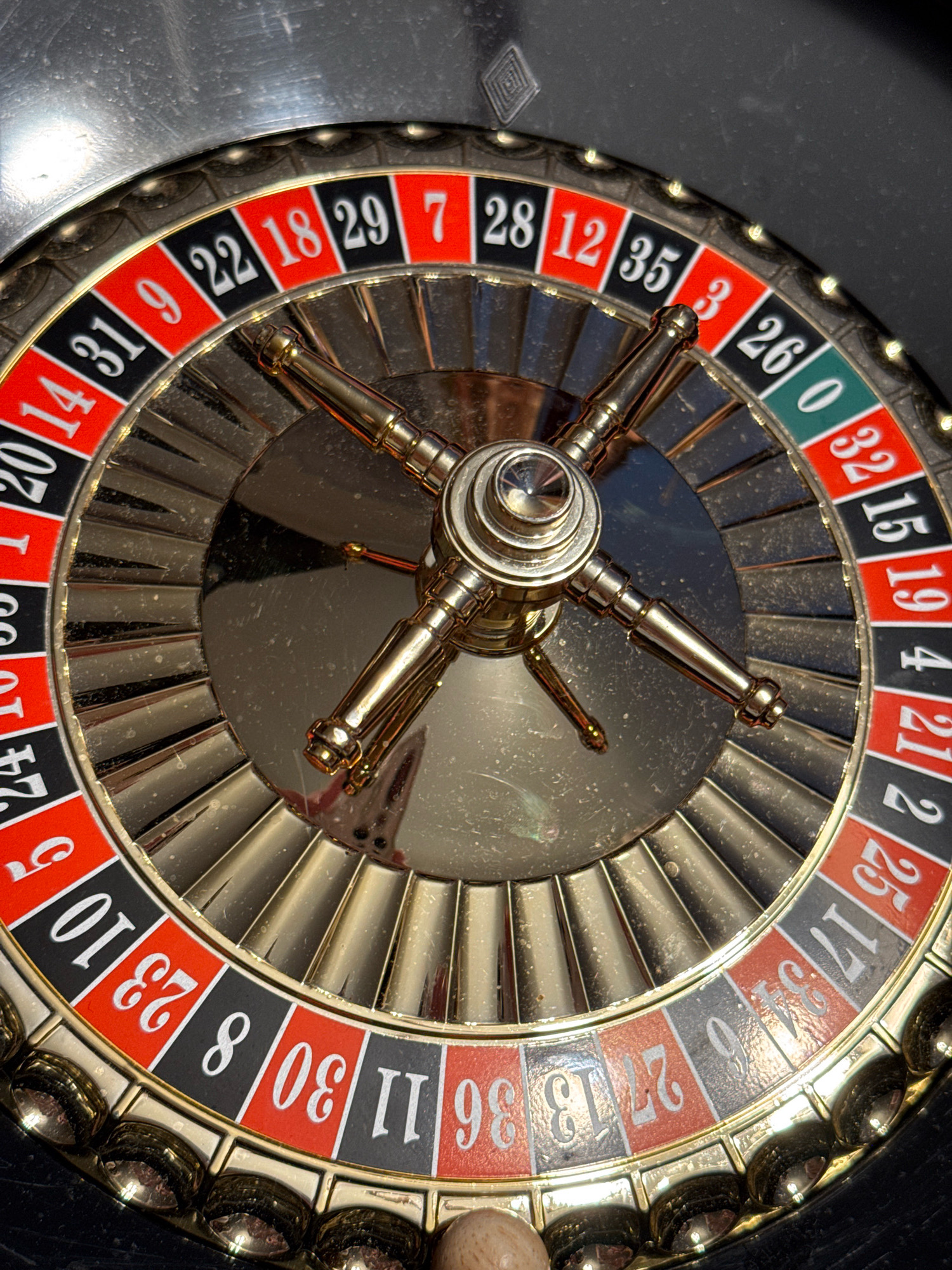 A close-up, overhead shot of a roulette wheel, positioned on a black surface, possibly a table. The roulette wheel itself is gold-colored with red and black numbered slots. The numbers visible include 14, 31, 9, 22, 18, 29, 7, 28, 12, 35, 3, 26, 0, 32, 15, 19, 4, 21, 2, 25, 17, 34, 6, 27, 13, 36, 30, 11, 8, 23, 10, and 5. The center of the wheel features a gold-colored metal mechanism with four spokes. The environment is dark with overhead lighting creating reflections on the metallic surfaces. There are no people or visible actions occurring. The image does not provide any information about the city of Saint Petersburg, Russia, or any specific location.