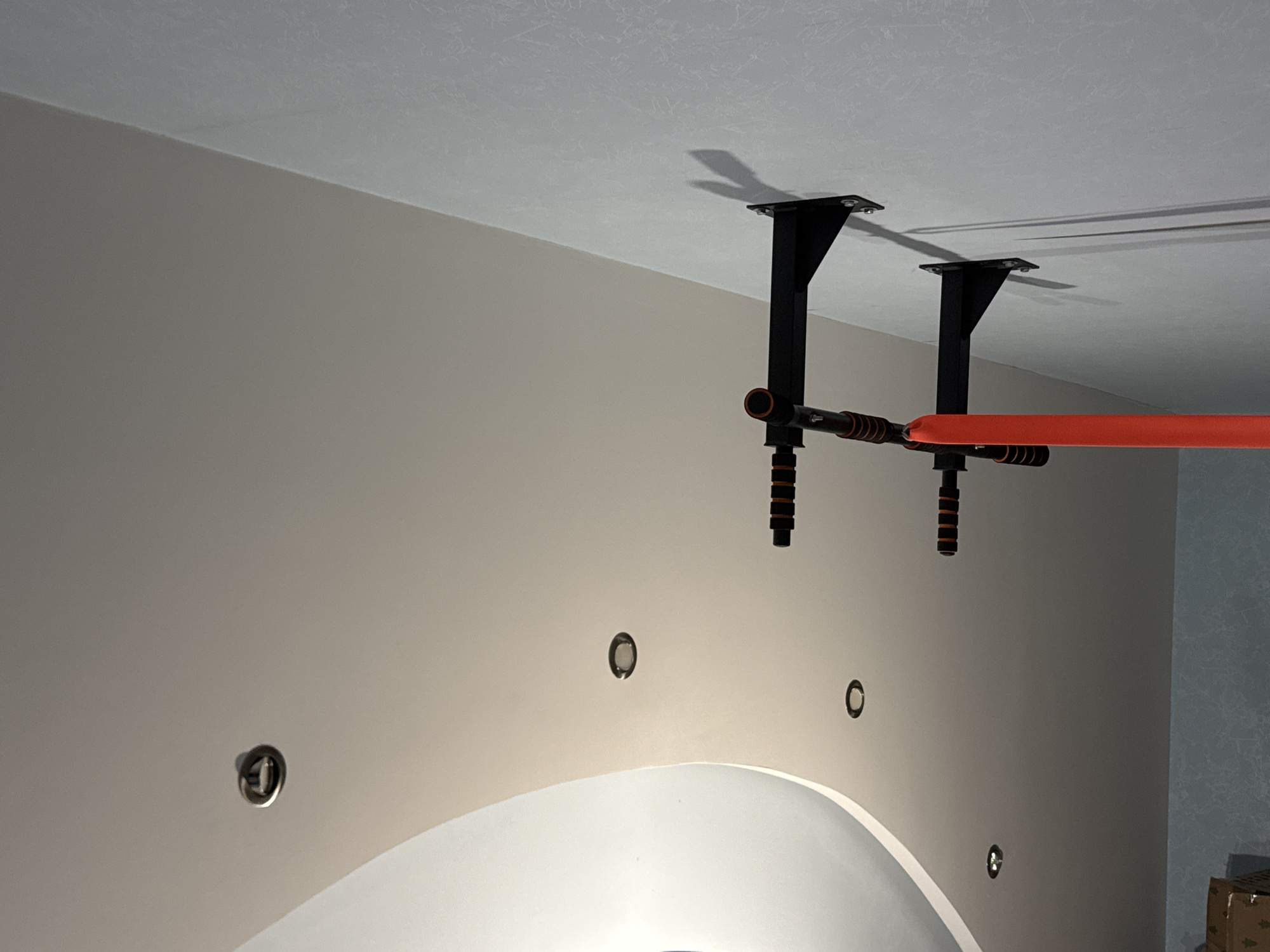 The image shows a wall-mounted pull-up bar in what appears to be an indoor setting. The pull-up bar is black with orange grips. Two recessed ceiling lights are visible on a curved ceiling above the wall. The wall itself has a textured pattern. The scene is devoid of people. The lighting suggests the photo was taken indoors, possibly in a home gym or recreational area. The location is unknown.