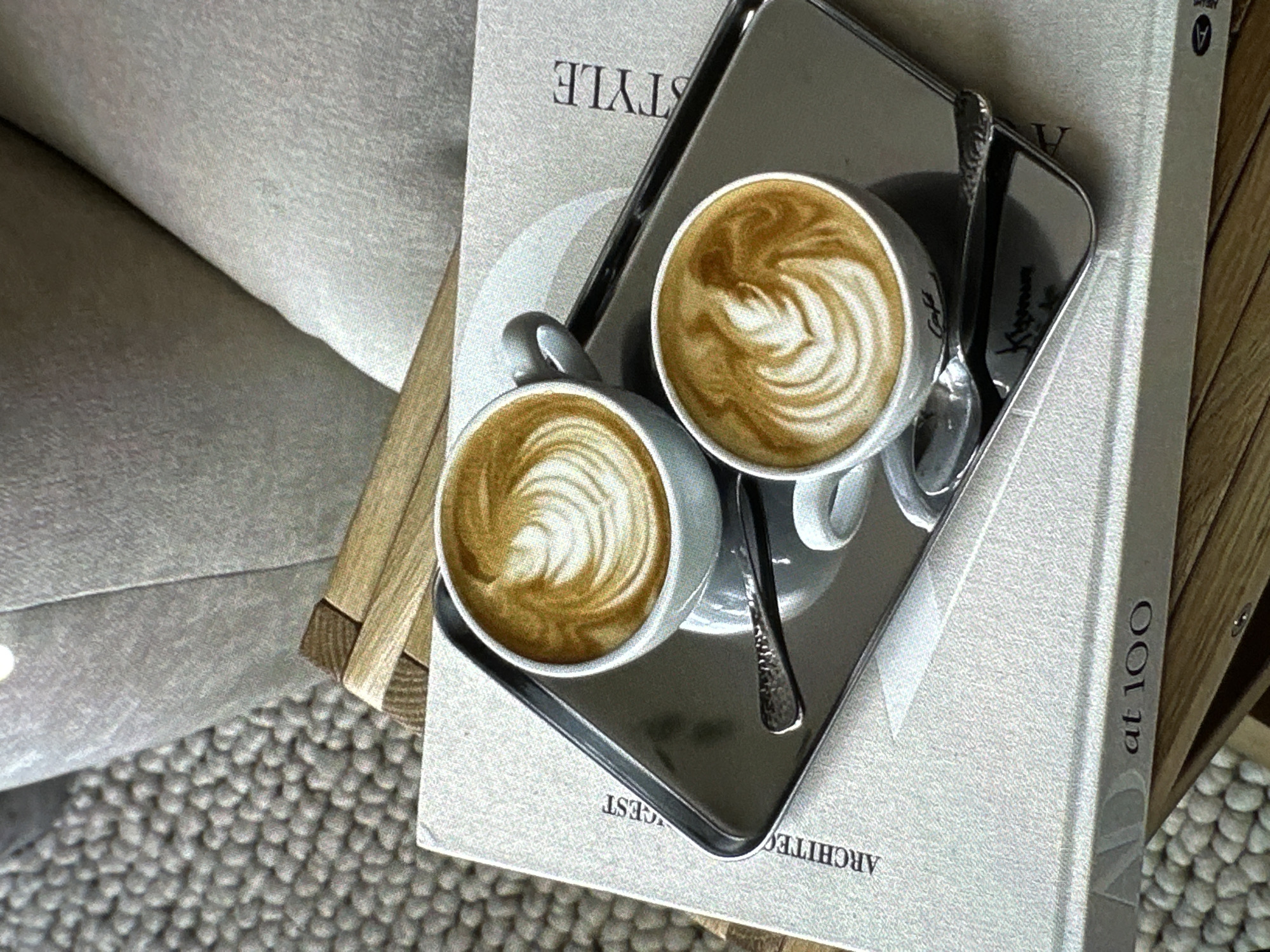 Two cups of latte art coffee on a silver tray with two spoons, placed on top of a book titled "Architectural Digest at 100: A Century of Style". The scene is likely inside a cafe or home. The environment consists of a couch/seat and rug with a pebble pattern. There is no indication of the location being Yerevan, Armenia.