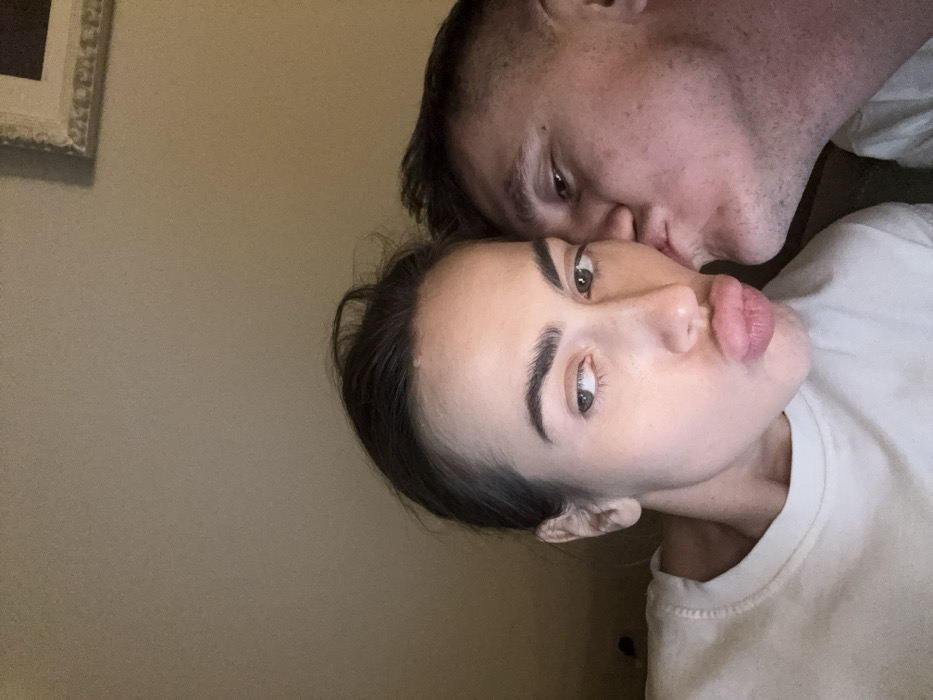 The scene depicts two individuals, one female and one male, in a close-up indoor setting in Venice, Italy. The female is positioned on the left, displaying dark hair pulled back, light skin, and her left eye looking to the right. Her lips are prominently pursed in a "duck face" expression. She wears a plain, light-colored crewneck garment. The male is on the right, with light skin and short dark hair. His right cheek is pressed against the female's right cheek, and his lips are puckered and in contact with her cheek, indicating a kiss. The background consists of a plain, light-beige wall. A fragment of a framed object with an ornate border is visible in the upper right corner. The lighting is soft and ambient, consistent with an interior space.