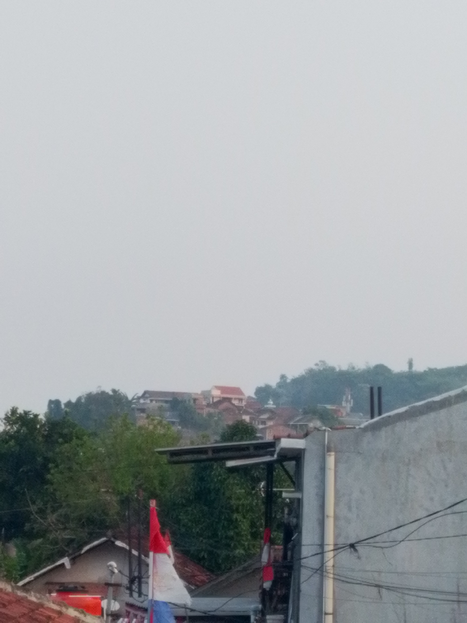 The image shows a view of a hillside residential area on an overcast day. In the foreground, the top of a grey, concrete building with electrical wires is visible. To the left, a portion of a smaller building with a red-tiled roof is seen, along with a white satellite dish. A red and white flag, likely the Indonesian flag, is partially visible and appears to be attached to a pole.
Further into the background, a dense cluster of trees obscures many of the houses that are nestled on the hillside. Some buildings with red-tiled roofs and white walls are discernible amidst the greenery. In the distance, a steeple of a church or similar structure can be spotted, along with other buildings that blend into the misty landscape. The overall impression is of a somewhat dense, hilly urban environment. The presence of the Indonesian flag suggests the location is in Indonesia, and the specific mention of Bandar Lampung points to that city as the setting.