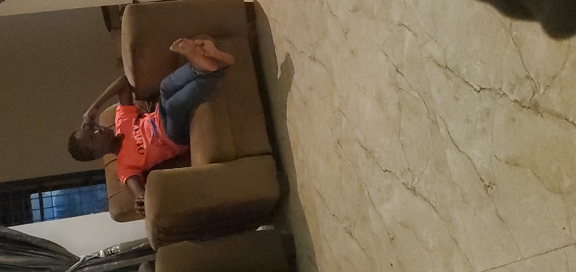 A young person, appearing to be of African descent, is seated on a brown sofa with their legs crossed and feet resting on the sofa arm. They are wearing an orange t-shirt with text and blue jeans. The person's right hand is touching their head, and they are looking to the side. The background shows a window with vertical bars, a curtain, and a wall. The floor is tiled with a marble pattern. The scene is indoors, suggesting a home environment. The lighting is dim, creating shadows.