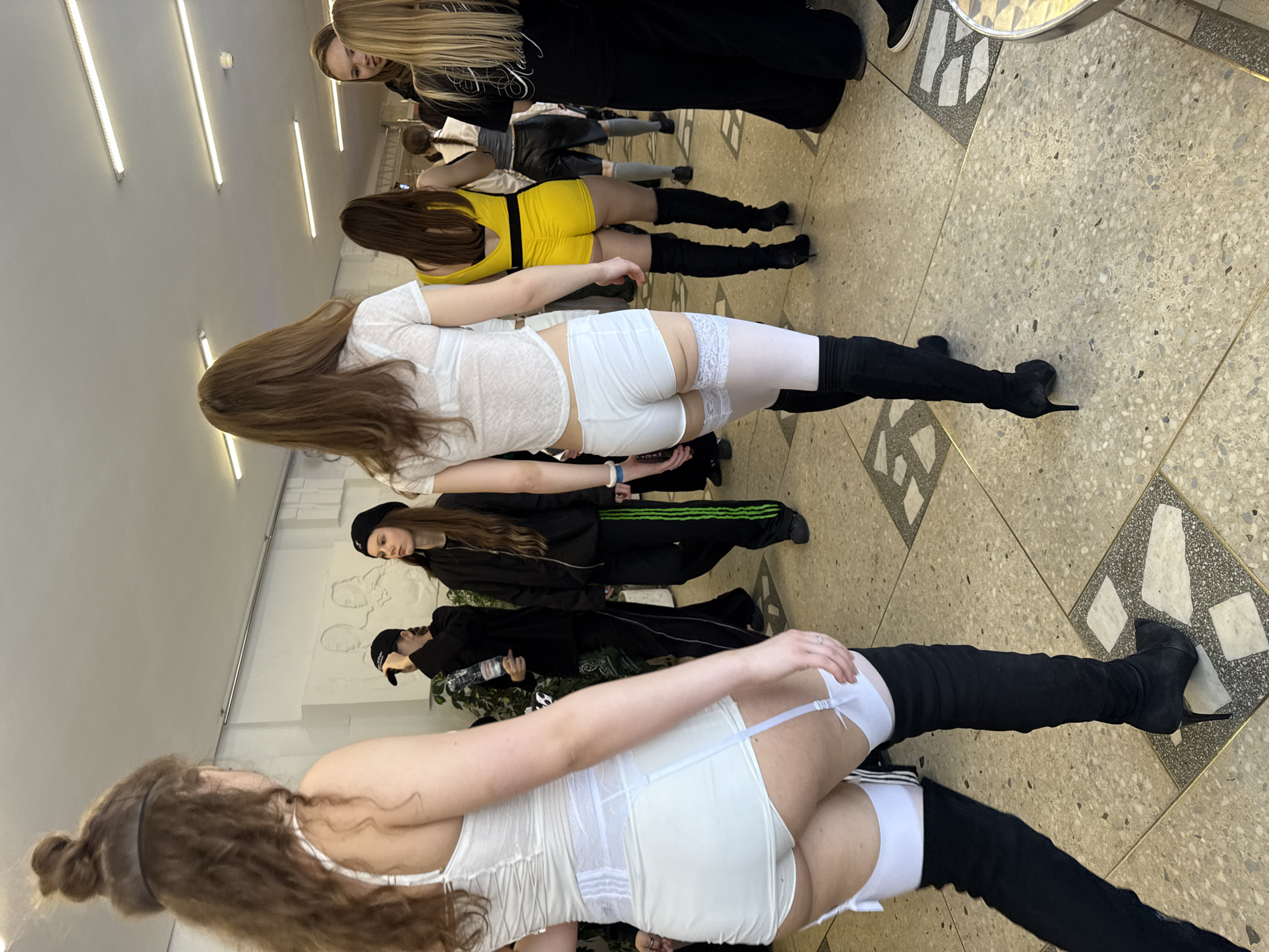 A group of women is standing in a hallway with white walls, fluorescent lighting, and a patterned tile floor. The women in the foreground are wearing white lingerie, white stockings, and black thigh-high boots. The woman in the middle wears a yellow dress and black boots. The rest are dressed in black clothing. The women are standing in a line and seem to be posing. Some people in the background are wearing black hats. The hallway is decorated with white relief sculptures. The location may be in the city of Perm, Russia, based on visual cues.