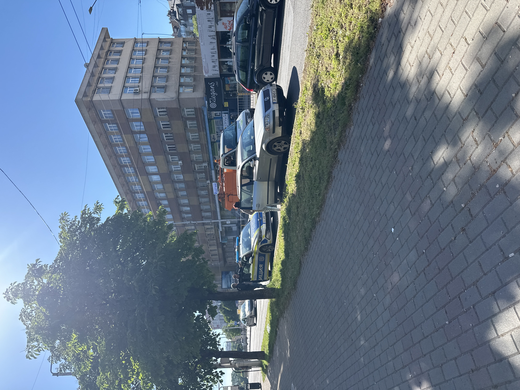 The image shows a street scene in Varna, Bulgaria. A multi-story building stands on the left. The sidewalk is paved with rectangular gray bricks. A row of parked cars lines the street, including a police car with Bulgarian "ПОЛИЦИЯ" markings visible. Two people are interacting near the police car. A tree stands between the viewer and the street. The sky is clear and blue. "Elegans" store is visible on the building next to a black car. A light pole and associated cables are also visible.