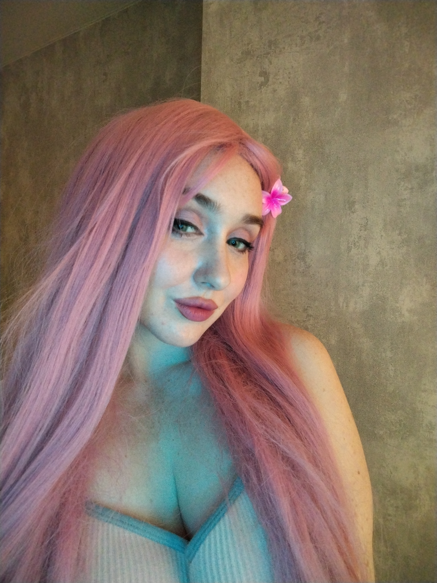 The image displays a medium close-up portrait of a woman with long, flowing, light pink hair, which cascades over her shoulders and chest. She is looking directly at the camera with a calm, slightly alluring expression, featuring a subtle smirk and light-colored eyes enhanced with eyeliner. Her eyebrows are well-defined, and she wears pink lipstick. A small, vibrant pink flower is tucked into her hair above her right ear.
She appears to be wearing a light-colored, possibly ribbed, top or bra with a blue trim, visible at the bottom of the frame. Her skin is fair, with some freckles visible on her shoulder and décolletage. The lighting casts a distinct blue-green hue over her chest and neck, while her face and hair retain warmer tones. The background is a simple, textured wall in shades of grey and brown, indicating an indoor setting. No specific activity beyond posing for the portrait is depicted. There is no visible text, and no cues for the time of day or weather are present.