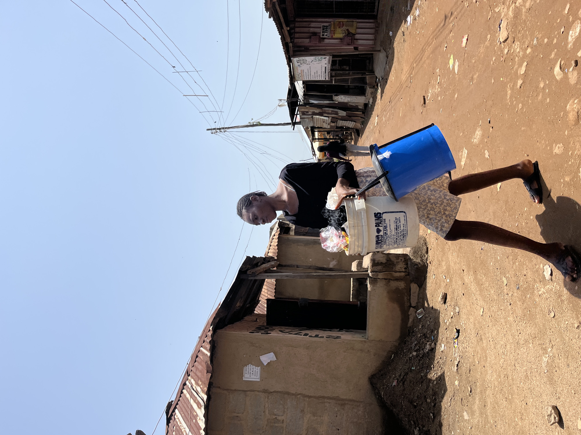 A young Black female is walking down a dirt road in the city of Jos, Nigeria, carrying two buckets. The female is wearing a black long-sleeved shirt, a floral skirt, and black sandals. One bucket is white and has "DIAMOND PAINTS" written on it. The other bucket is blue. She is walking towards the right side of the image. The background consists of buildings with corrugated iron roofs, some of which appear to be makeshift structures or shops. There are also power lines running overhead. The ground is dusty and scattered with debris. Another person, likely a female, is walking further down the road in the same direction as the main subject. The sky is clear and blue, suggesting a sunny day. The overall impression is of a scene from an informal settlement or market area in Jos.