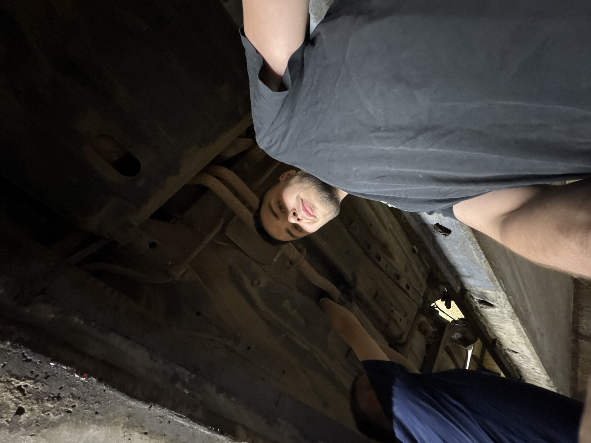 The image depicts two men from a low-angle perspective, positioned beneath the heavily rusted undercarriage of a vehicle, likely within an automotive repair pit or garage. The overall lighting is dim and artificial.
On the right side of the frame, a man with short dark hair and a beard looks directly at the camera, his expression neutral with a slight smile. He is wearing a dark gray or black t-shirt, and his right arm is partially visible.
On the left, partially obscured, a second man's right arm, clothed in a dark blue t-shirt, is extended upwards, appearing to be interacting with or inspecting a component of the vehicle's underside. His face is not visible.
Above the men, the underbody of the vehicle reveals extensive rust across its metallic surfaces, including visible exhaust pipes, frame rails, and various underbody panels. Some areas show accumulated dirt and grime. A small, dark, oblong hole is present in one of the underbody panels on the upper right side of the frame.
The immediate environment visible includes parts of a distressed concrete or heavily soiled metallic structure on the far left and in the lower foreground, consistent with the edge of a workshop pit or a vehicle lift support.
The image itself does not contain any visual information to determine its specific geographical location, including the city of Perm, Russia.