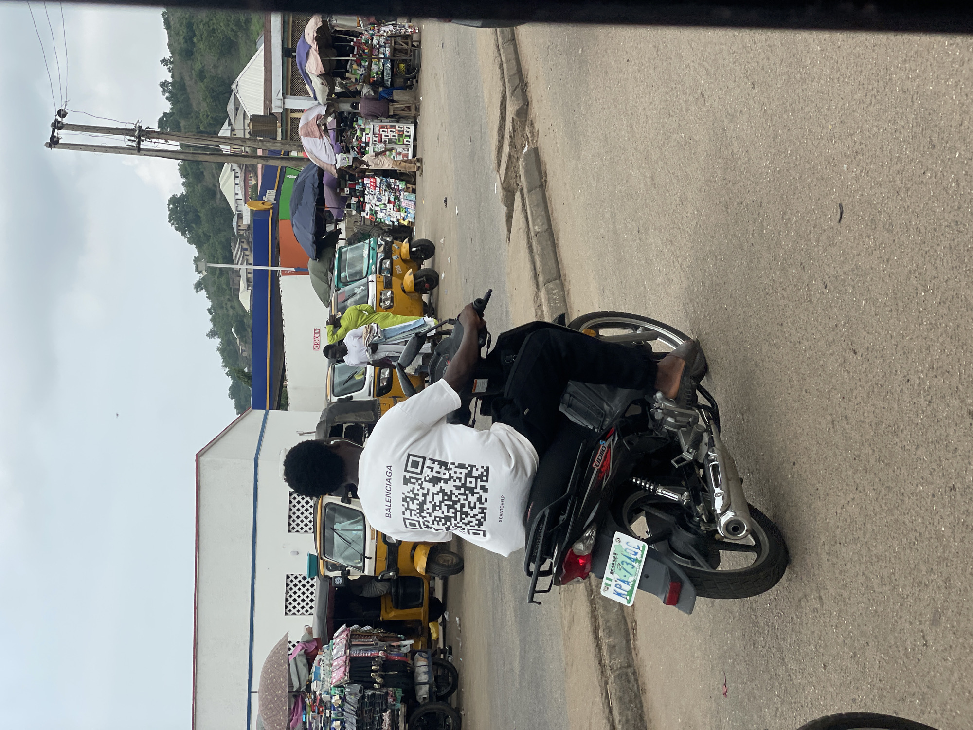A person is riding a black motorcycle on a paved street in Lokoja, Nigeria. The rider is wearing a white t-shirt with a large QR code and the word "BALENCIAGA" printed on the back, along with the text "SCANTOWELP". The rider is also wearing black pants and dark shoes. The motorcycle has a green license plate that reads "KGI KPA 7340C". In the background, there are several yellow auto-rickshaws, market stalls with umbrellas, buildings, and a hillside with scattered houses. The sky is overcast.