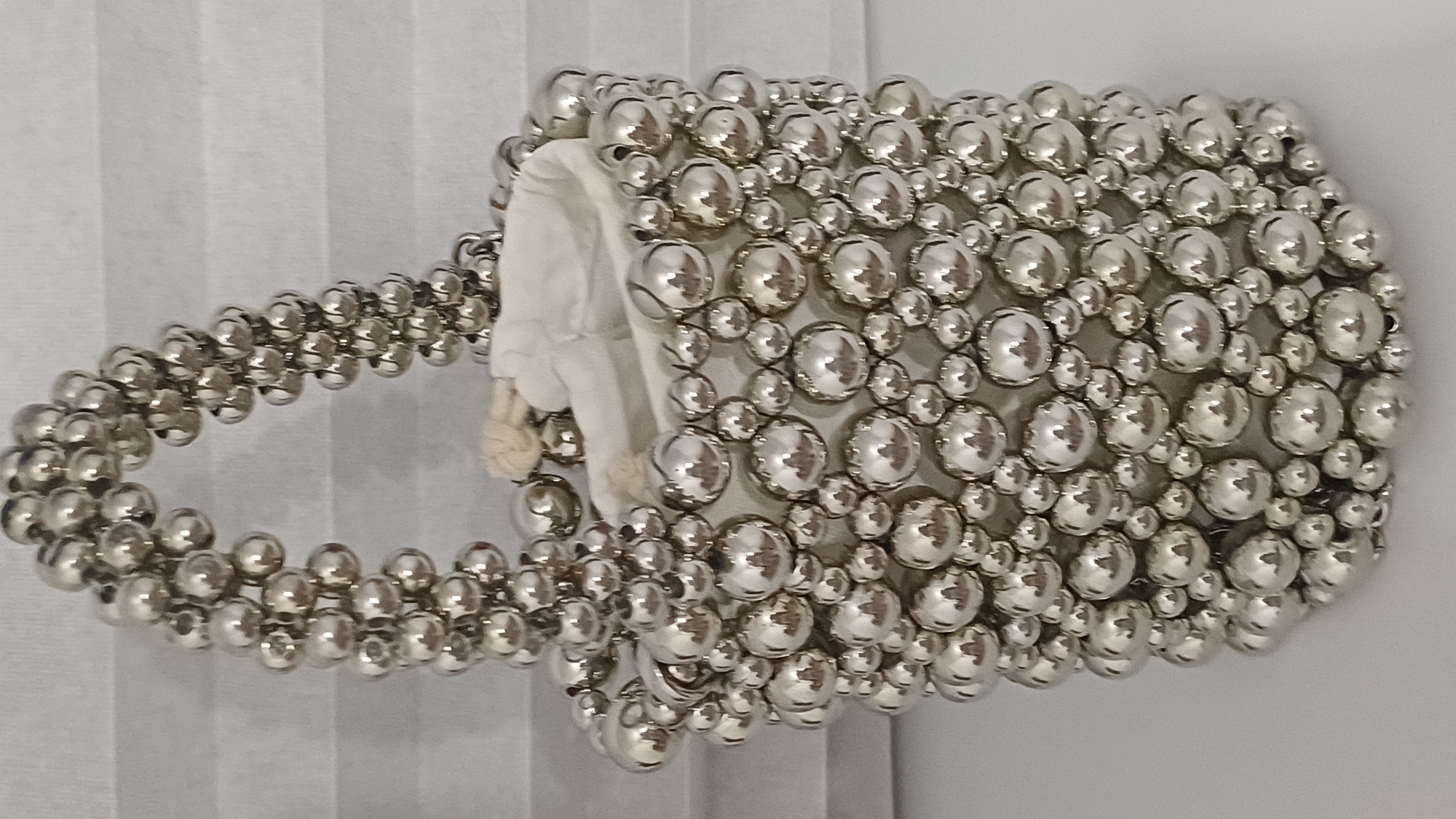 A close-up, eye-level, vertical shot shows a beaded handbag with a white interior on a white surface. The handbag is covered in numerous small, silver-colored, spherical beads of varying sizes, creating a textured, metallic surface. A white drawstring closure with a small loop is visible at the top of the bag, peeking out from within. The handle of the bag is also constructed from the same silver beads, forming a loop. The background is a neutral, light gray, horizontally pleated surface. The lighting is soft and even, with some reflections visible on the shiny beads. There are no people or discernible activity in the image, and the location is unknown.