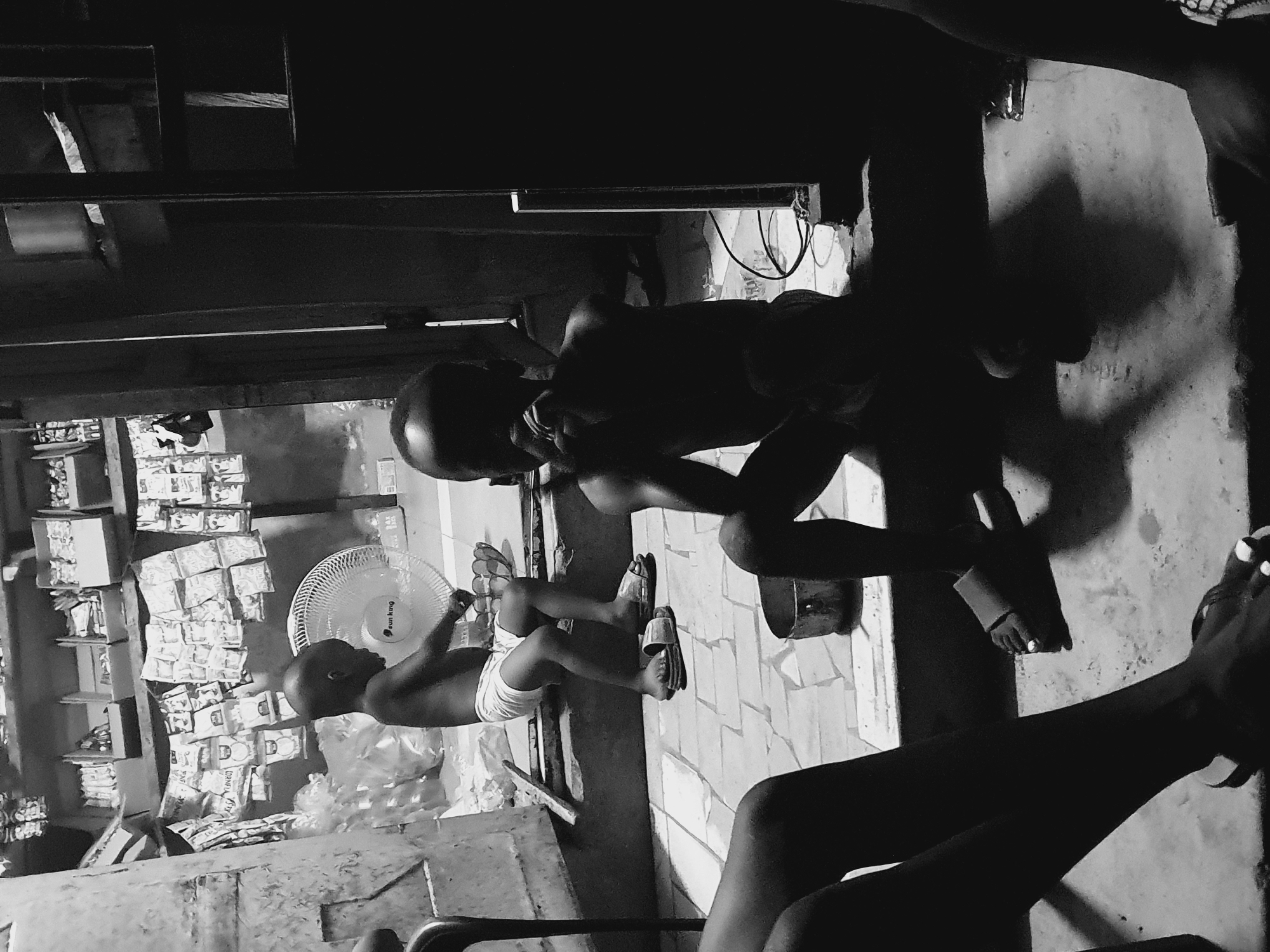 A black and white photograph shows an interior scene in Lokoja, Nigeria. In the lower left, shelves stocked with packaged goods are visible. A young child wearing underwear and sandals is playing with a fan in front of the shelves. A person bends low, seemingly talking to the child. Parts of other people, including their legs and feet, are visible. The scene is dimly lit. The floor appears to be tiled. The environment looks like a small store or living area in Lokoja, Nigeria.