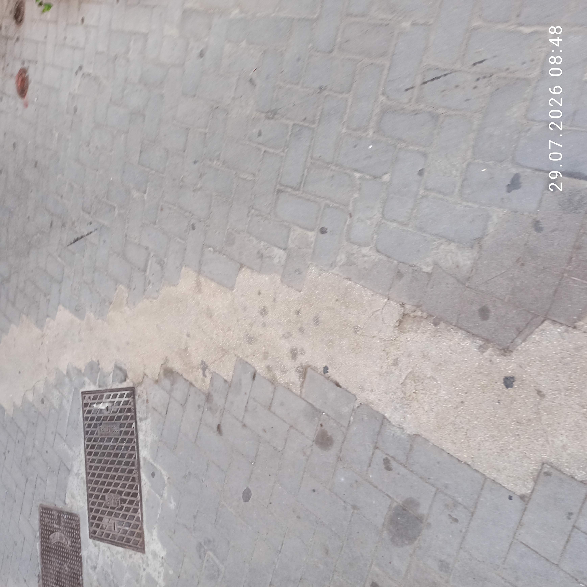 The image shows a textured, gray, paver-stone sidewalk with a lighter, sandy-colored strip running diagonally across it. Two metal grate covers are visible on the left side of the sidewalk. One grate is rectangular and features a diamond pattern with two rectangular indentations. The second, partially visible grate is also rectangular and displays a circular emblem within its diamond pattern. The sidewalk appears to be somewhat damp in the lighter colored section. The timestamp "29.07.2026 08:48" is visible in the lower right corner of the image. The scene is an outdoor urban environment.