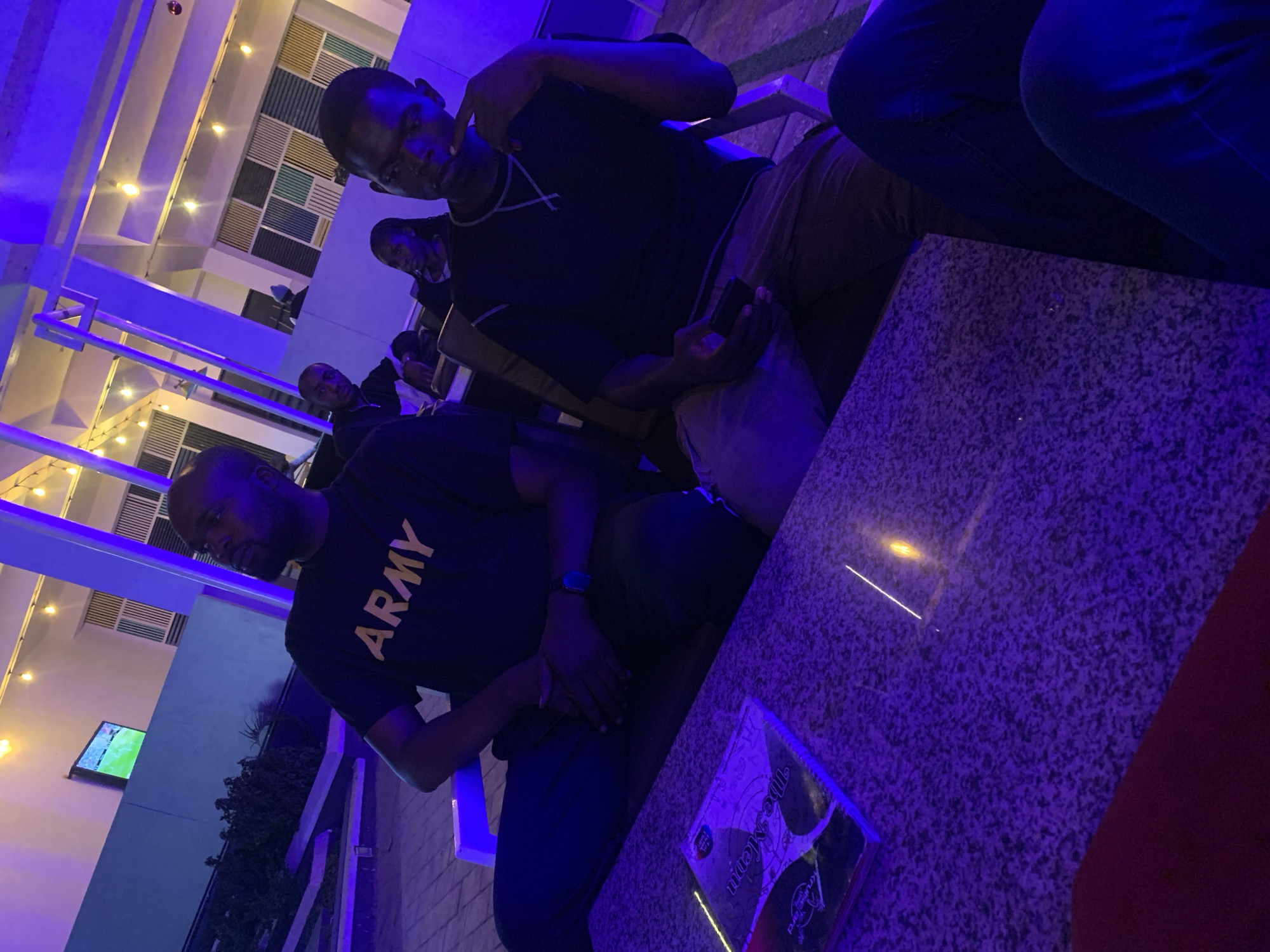 A group of men are seated at an outdoor table under blue-tinted lighting. One man wears a dark-colored "ARMY" t-shirt. Another man sits opposite him, holding a phone and making a gesture with his hand. Other people are partially visible in the background. A menu is on the table. The setting appears to be an outdoor dining area in Gura Top, Nigeria.
