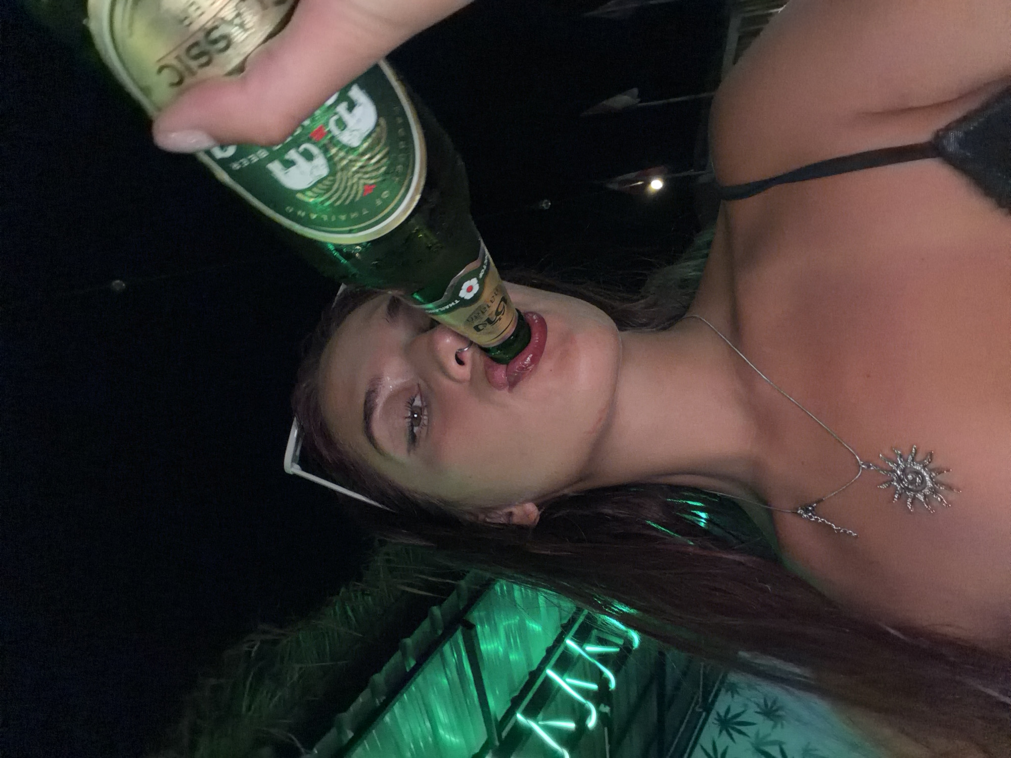A young woman with long, dark hair and light-colored framed glasses perched on her head is captured in a close-up, low-angle shot, drinking from a green bottle of beer. She has a sun-shaped pendant necklace resting on her chest, and her eyes are slightly closed as she takes a sip. The background is dark, with blurry green lights and foliage visible, suggesting an outdoor setting at night. The presence of the beer bottle and the relaxed posture indicate a casual social occasion. The location is identified as Karon, Thailand, though specific landmarks are not evident in the image itself.