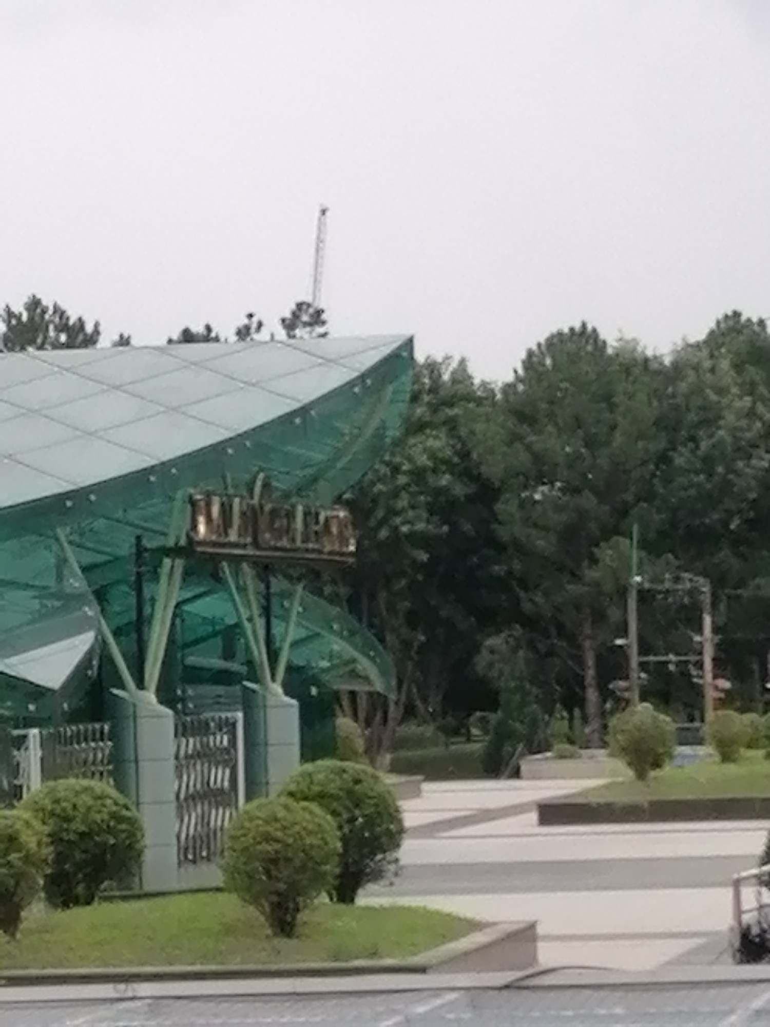The image shows a wide shot of an outdoor area with a modern, glass-roofed structure in the foreground. The structure has a curved, green, translucent roof supported by visible white pillars. A sign with metallic lettering, partially obscured, is mounted on a dark horizontal beam beneath the roof. To the left of the structure, a decorative metal gate is visible between two concrete pillars. Surrounding the structure are manicured bushes and grass. In the background, a dense line of mature trees forms a backdrop. Above the trees, a tall, thin structure, possibly a crane, is visible against a bright, overcast sky. The ground in front of the structure appears to be paved with light-colored stones or tiles, arranged in a patterned walkway. The scene suggests a public space or park in Da Lat, Vietnam.