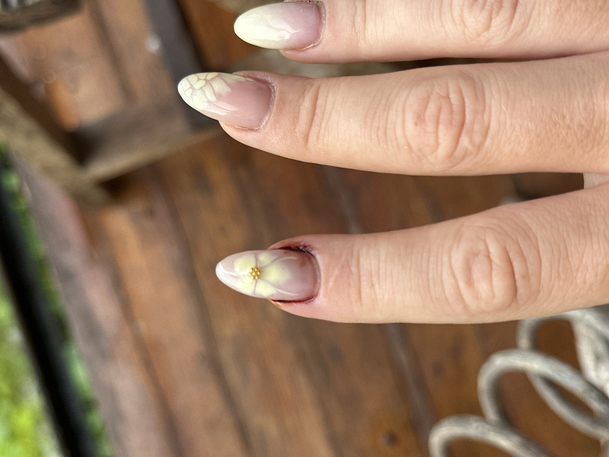 A close-up shot focuses on the fingernails of a person's left hand, which has an oval-shaped manicure with a pale yellow floral design and a small gold bead at the center of the flower. The nails are resting on a wooden surface, likely a deck or table, in an outdoor setting. The background is blurred, showing greenery and wooden structures. The person's hand is positioned in front of a set of grey metal railings, which are also out of focus. The lighting suggests it is daytime. The image does not contain any identifiable landmarks or contextual clues to pinpoint its location in North Kuta, Indonesia.