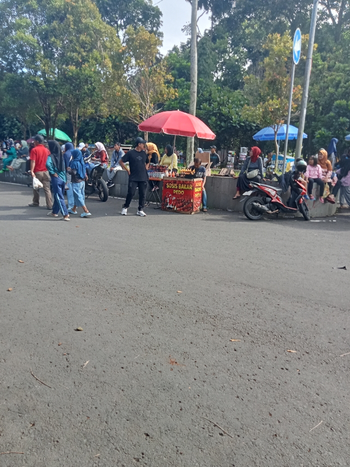 This image depicts a bustling outdoor scene in Singaparna, Indonesia, likely a street market or a public gathering area adjacent to a park. In the center, a vendor operates a food stall under a bright red umbrella. Several people, including adults and children, are gathered around this stall, some standing and observing, while others appear to be interacting with the vendor.
The vendor's stall prominently displays the text "SOSIS BAKAR PEDO" and "SOSIS BAKAR PEDAS," indicating that grilled sausages, including spicy varieties, are being sold. Various skewered food items are visible on display.
To the left of the stall, a group of people, many wearing headscarves (hijabs), are either walking or standing together. To the right, two motorcycles are parked near a low concrete wall where several individuals, including children, are seated. Above this area, a blue circular road sign with a white directional arrow pointing left is visible.
The background is filled with numerous green trees, suggesting a park or a heavily vegetated area. Other smaller green and blue umbrellas can be seen further back, possibly indicating more stalls or shaded seating. The foreground shows a wide, paved asphalt road, appearing relatively clear of traffic but serving as a space for pedestrians. The scene is illuminated by natural daylight, suggesting it is a clear day, possibly late morning or afternoon, with a casual and active atmosphere.