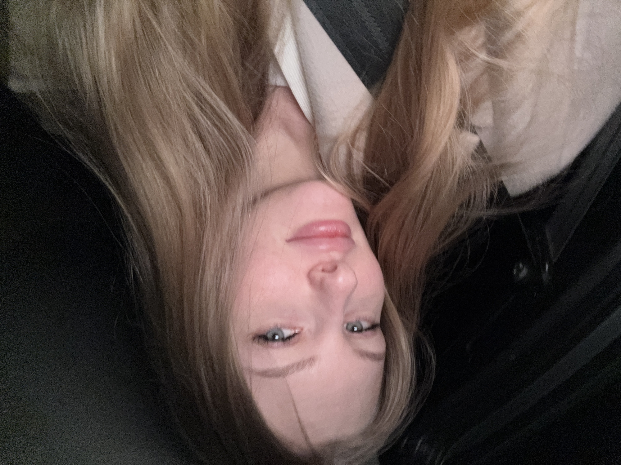 The image is an upside-down close-up portrait of a young woman with long, flowing light brown hair. In a right-side-up orientation, her face is the central focus, featuring pale skin, light blue eyes with a soft, upward gaze, and subtly defined eyebrows. Her pink lips are gently parted in a faint, serene smile, conveying a sense of calm and contentment.
She appears to be wearing a light-colored collared top, possibly white or cream, visible at her neck, beneath a darker garment that could be black or dark grey. The background is predominantly dark and indistinct, suggesting an indoor environment or low-light conditions. Some of the cropped views hint at textured material, possibly fabric or upholstery, which could indicate a setting like a car interior or a softly lit room. The lighting is soft, illuminating her face and hair. There is no visible text in the image.