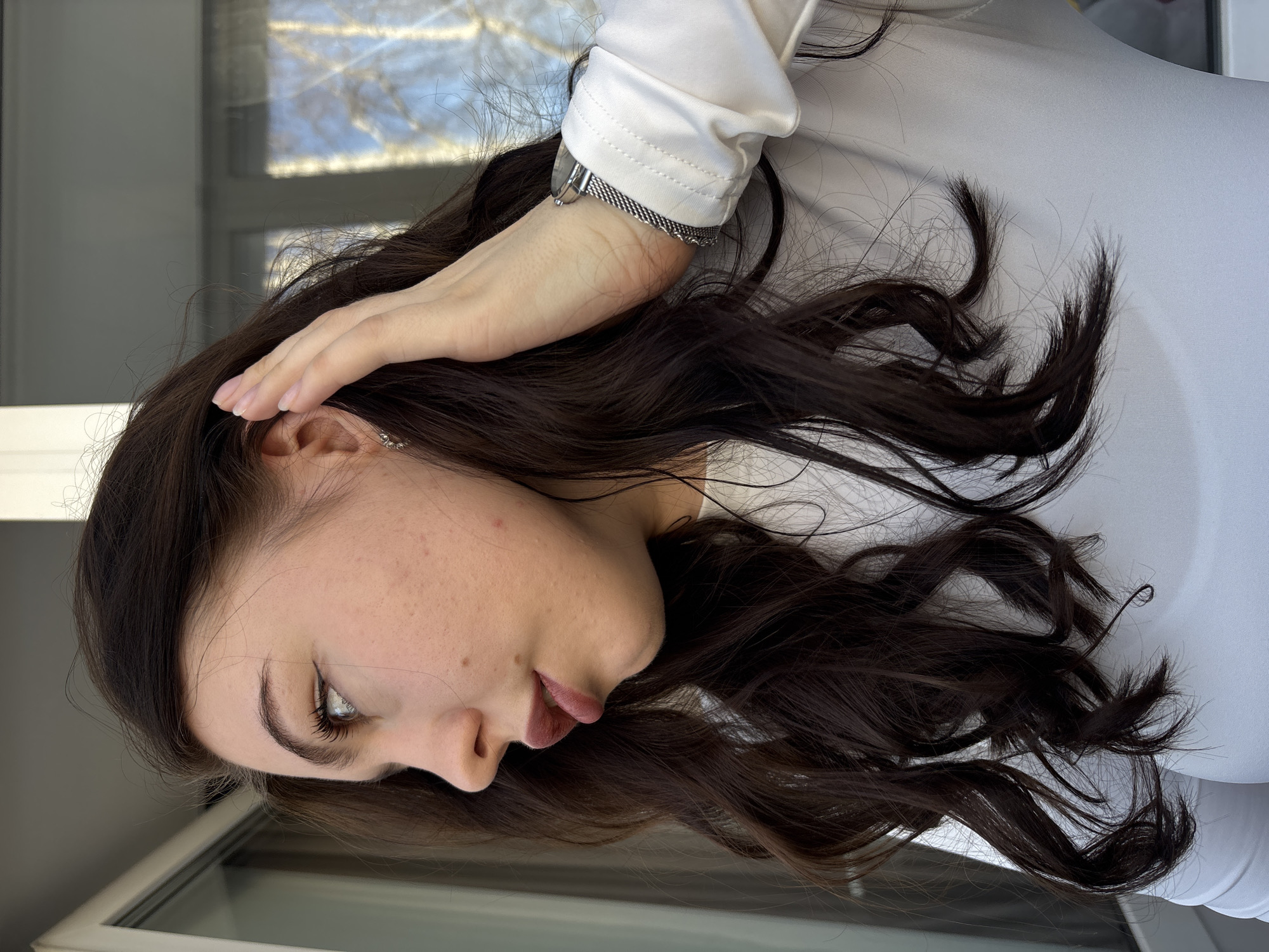 A close-up, right-profile shot of a female in the city of Perm, Russia. She has long, wavy dark brown hair and fair skin with minor blemishes. Her right eye features dark eyeliner, and her lips are painted a dark pink/mauve. She is wearing a white long-sleeved top. A silver-toned mesh-strap watch is present on her left wrist, and a small silver hoop earring is visible in her right ear. Her left hand is raised, with fingers lightly touching the hair near her right ear. Her gaze is directed upwards and to the left. The background shows a white window or balcony door frame, with a bright blue sky and bare tree branches visible through the glass, indicating an outdoor daytime setting.