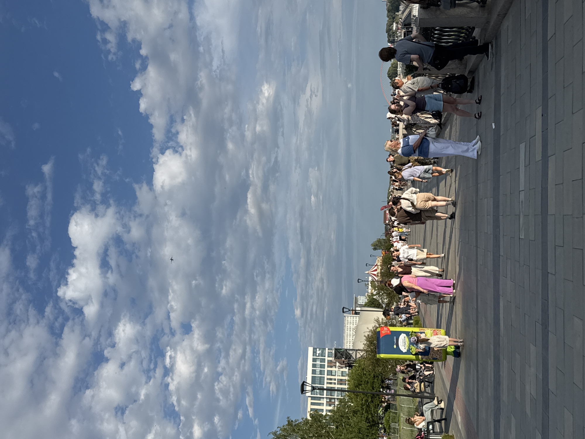 An aerial view of the embankment in Novosibirsk, Russia, shows a wide, paved walkway bustling with pedestrians under a partly cloudy sky. In the distance, a modern building with large windows and a Ferris wheel can be seen. A small airplane is visible in the sky. People are strolling along the embankment, some sitting on benches, while others are gathered in small groups. The sun is casting long shadows, indicating it is late afternoon or early evening. The overall atmosphere is one of leisure and recreation.