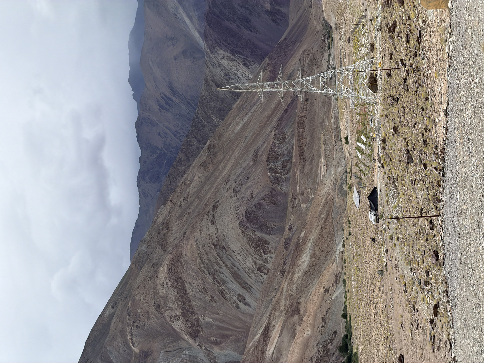 The image depicts a vast, arid mountainous landscape under a cloudy sky. In the foreground, a rough, rocky terrain is visible, leading into a flatter, stony expanse dotted with sparse dry vegetation. A white electrical pylon stands prominently in the mid-ground, its lattice structure stark against the muted tones of the surroundings. To the left of the pylon, a cluster of small, dark structures are present, possibly shelters or small buildings, with a white vehicle parked nearby. Further into the landscape, more distant, rugged mountains rise, their slopes layered with various shades of brown and gray. The scene appears to be located in Kiari, India, a region known for its high-altitude deserts and dramatic geological formations.
