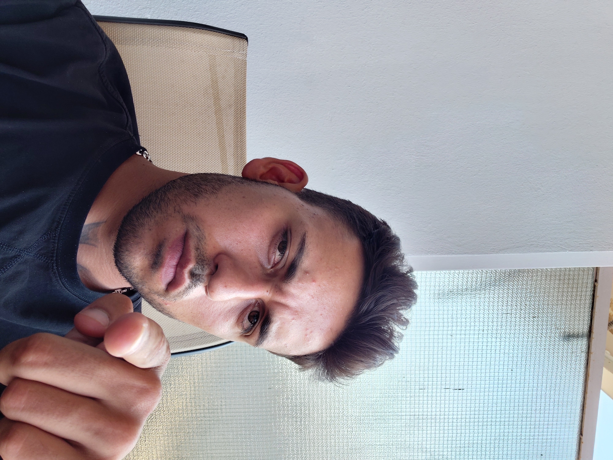 The image is a close-up selfie of a young man with dark hair and a beard, wearing a black t-shirt and a chain necklace. He is sitting in a chair with a tan fabric seat, in front of a white wall. To his right, there is a window with textured glass. The man is looking directly at the camera and pointing his right index finger forward, towards the viewer. The scene appears to be indoors, possibly in a residential or office setting. Based on the general architectural style and the light, it is plausible that this scene is located in Municipal Unit of Aegio, Greece, a coastal area in the Peloponnese.