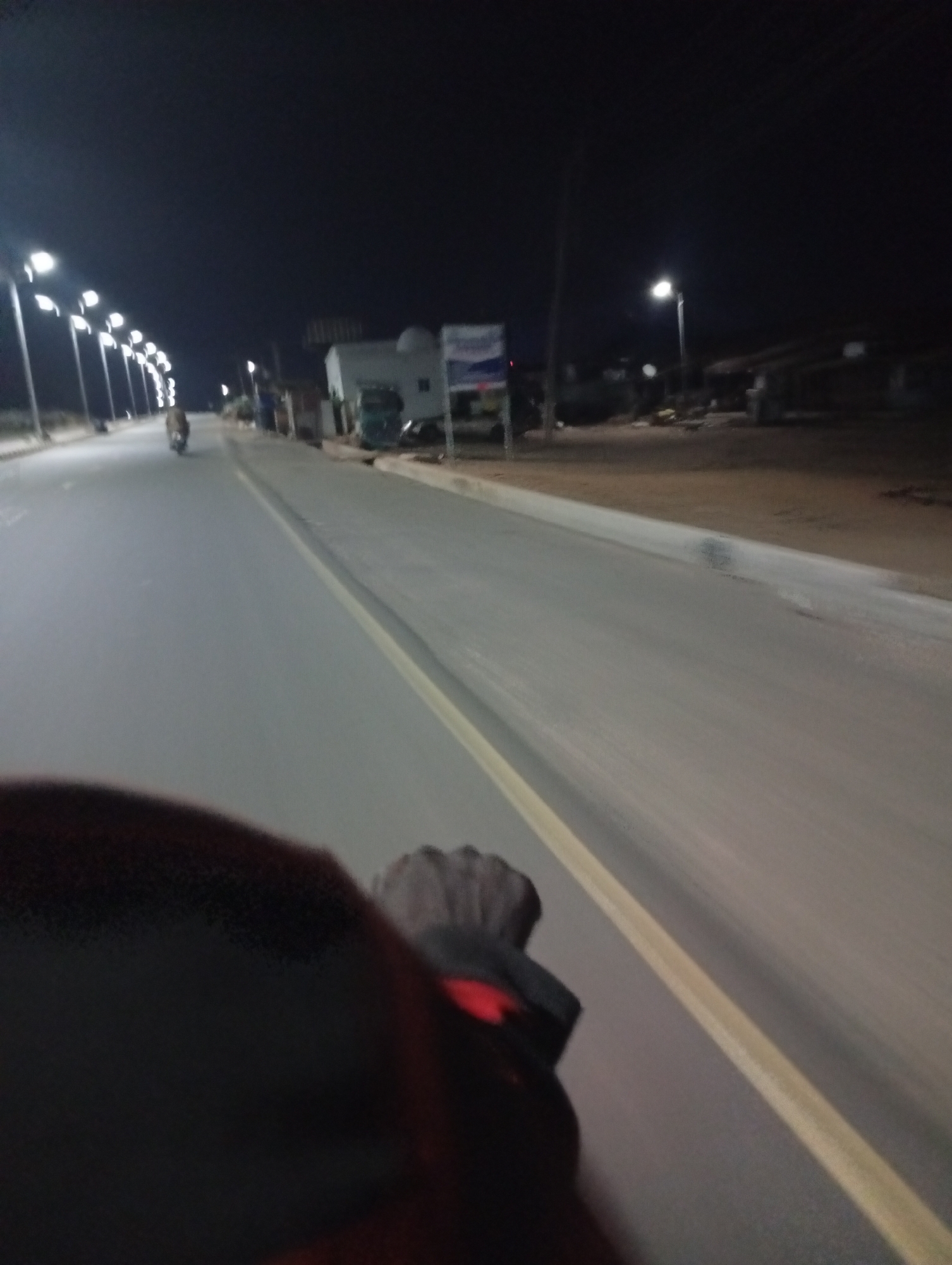 A nighttime view from what appears to be a moving vehicle on a paved road in Adankolo, Nigeria. The road is lit by streetlights that line one side of the road and is divided by a yellow line. In the distance, another vehicle (likely a motorcycle) is visible. The foreground shows a hand gripping the handlebar of the moving vehicle. Some buildings are visible to the side of the road.
