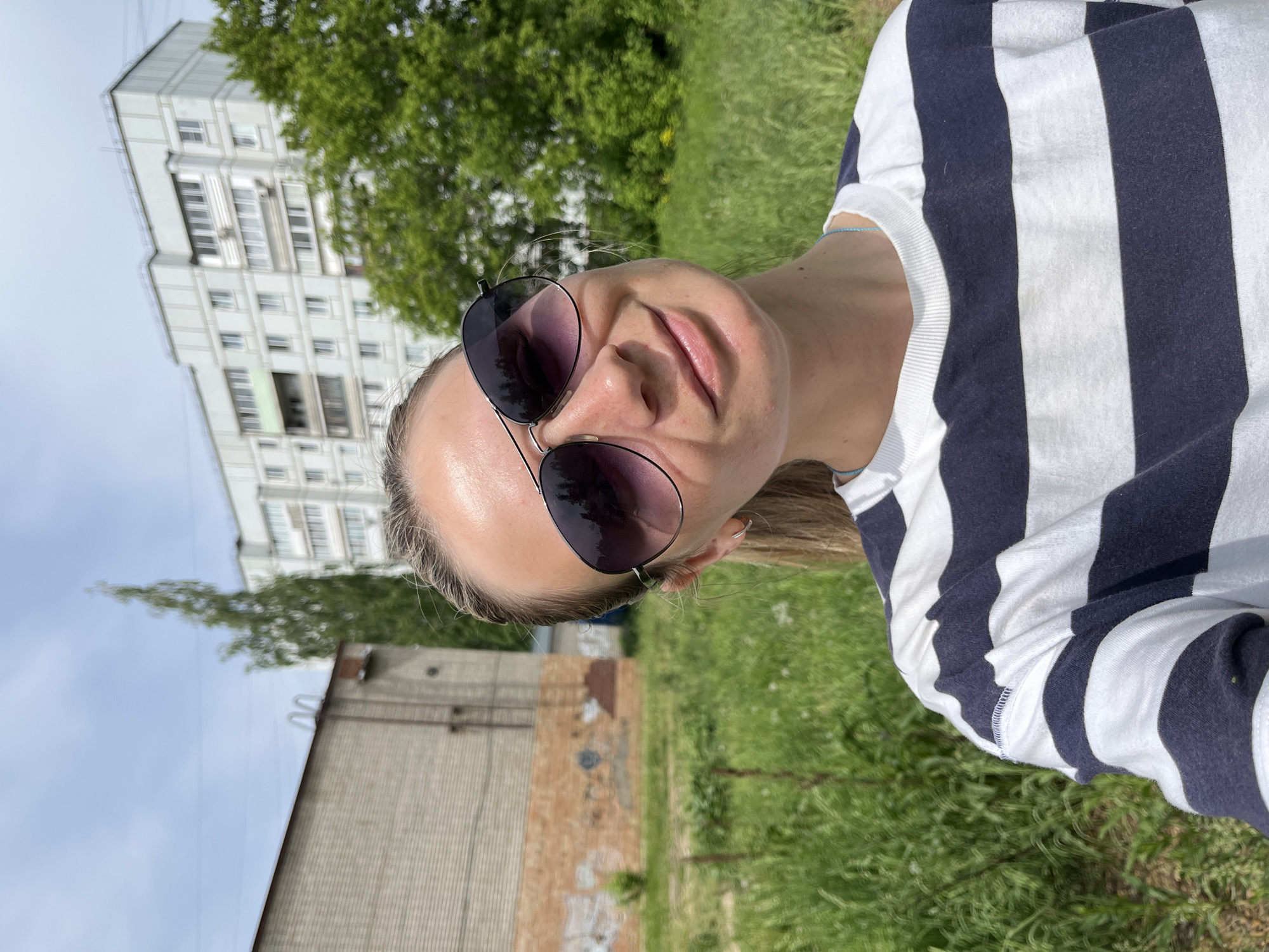 A woman with blonde hair and sunglasses is taking a selfie outdoors. She is wearing a white and navy striped top. Behind her is a grassy area and a building with a gray facade and brick accents. Further in the background, a multi-story apartment building is visible. The location is identified as Chaikovsky, Russia, based on available data.