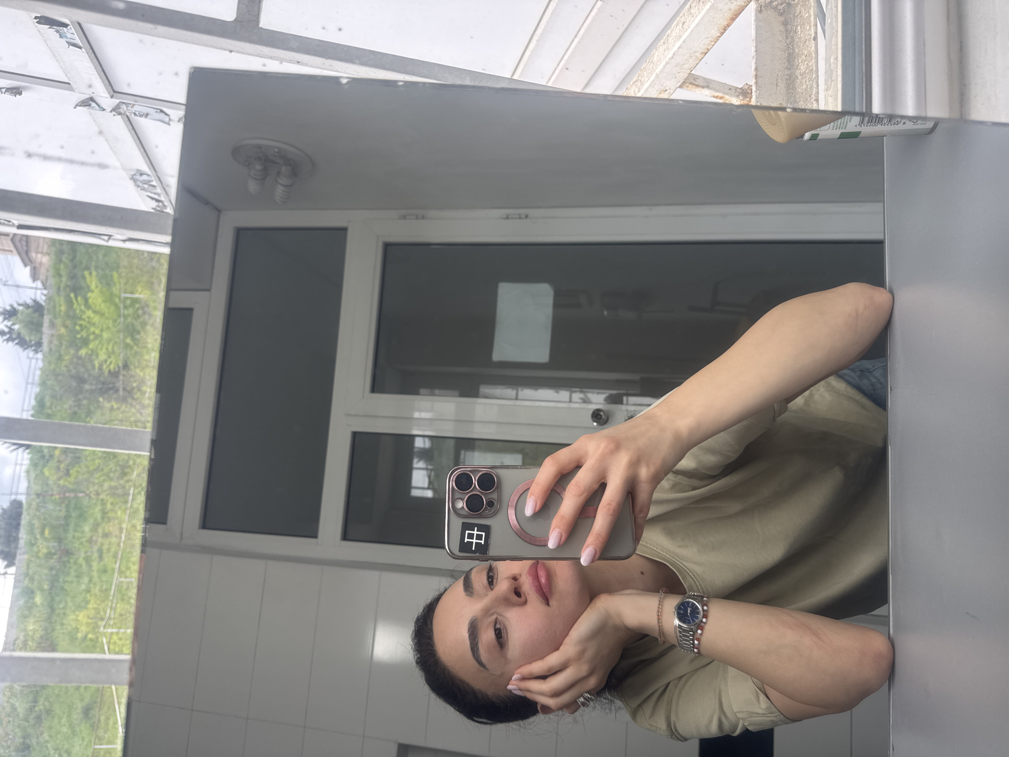 A woman takes a selfie in a mirror. She holds a phone in her right hand, its case decorated with a Chinese character sticker. She leans her head against her left hand. A watch is visible on her left wrist. The reflection in the mirror reveals a white-tiled wall, a window showing greenery, and a door leading to what appears to be an exterior covered area. The location is Tbilisi, Georgia.