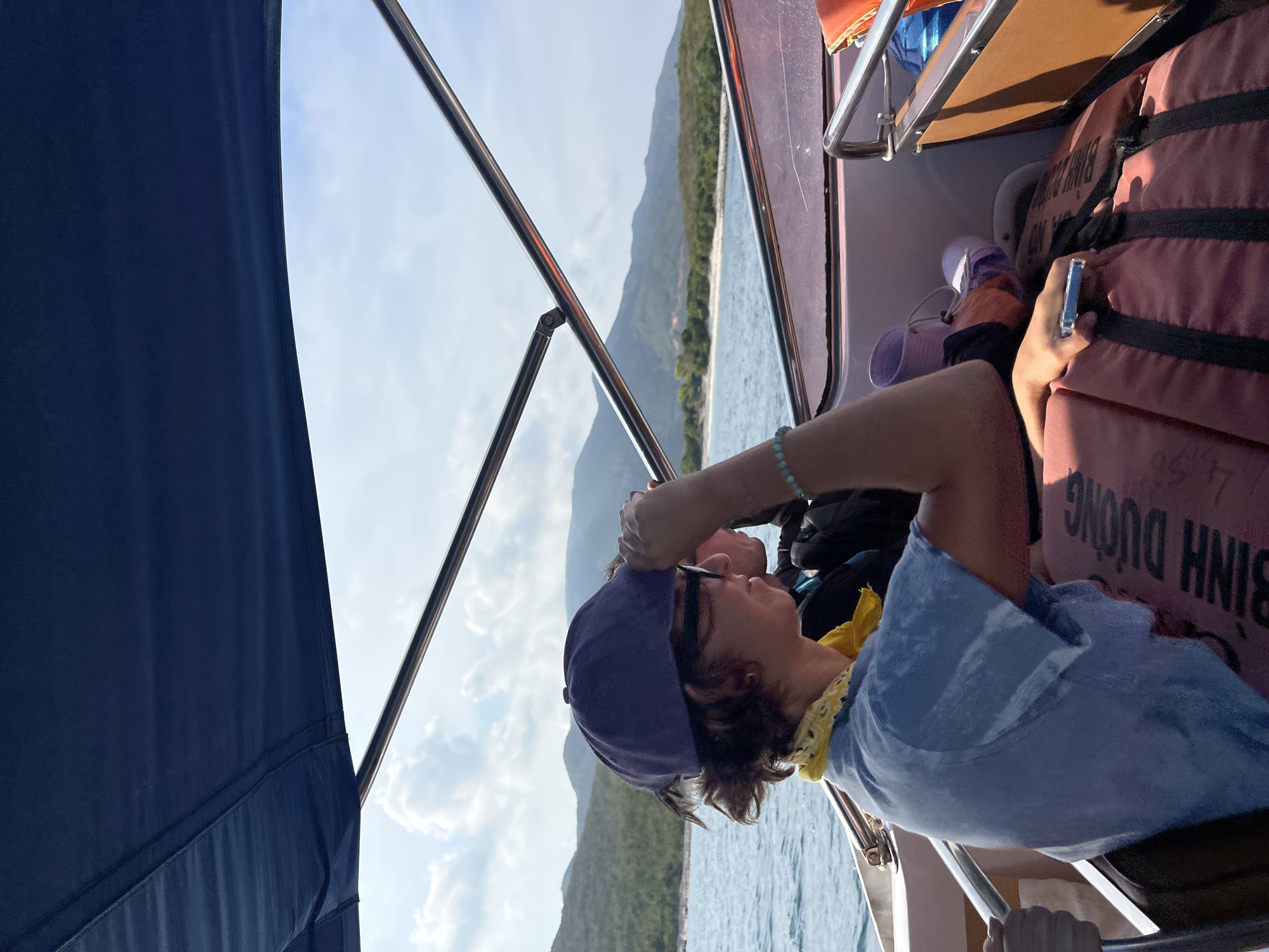 A sideways outdoor shot from a boat in Bac Nha Trang Ward, Vietnam, shows a person wearing a blue shirt and a baseball cap looking at their cell phone. The person is sitting next to a stack of orange life jackets. In the background, there are mountains and the ocean. The sky is blue with some clouds. The shot is taken from inside the boat and includes parts of the boat's structure such as the awning and metal railing.